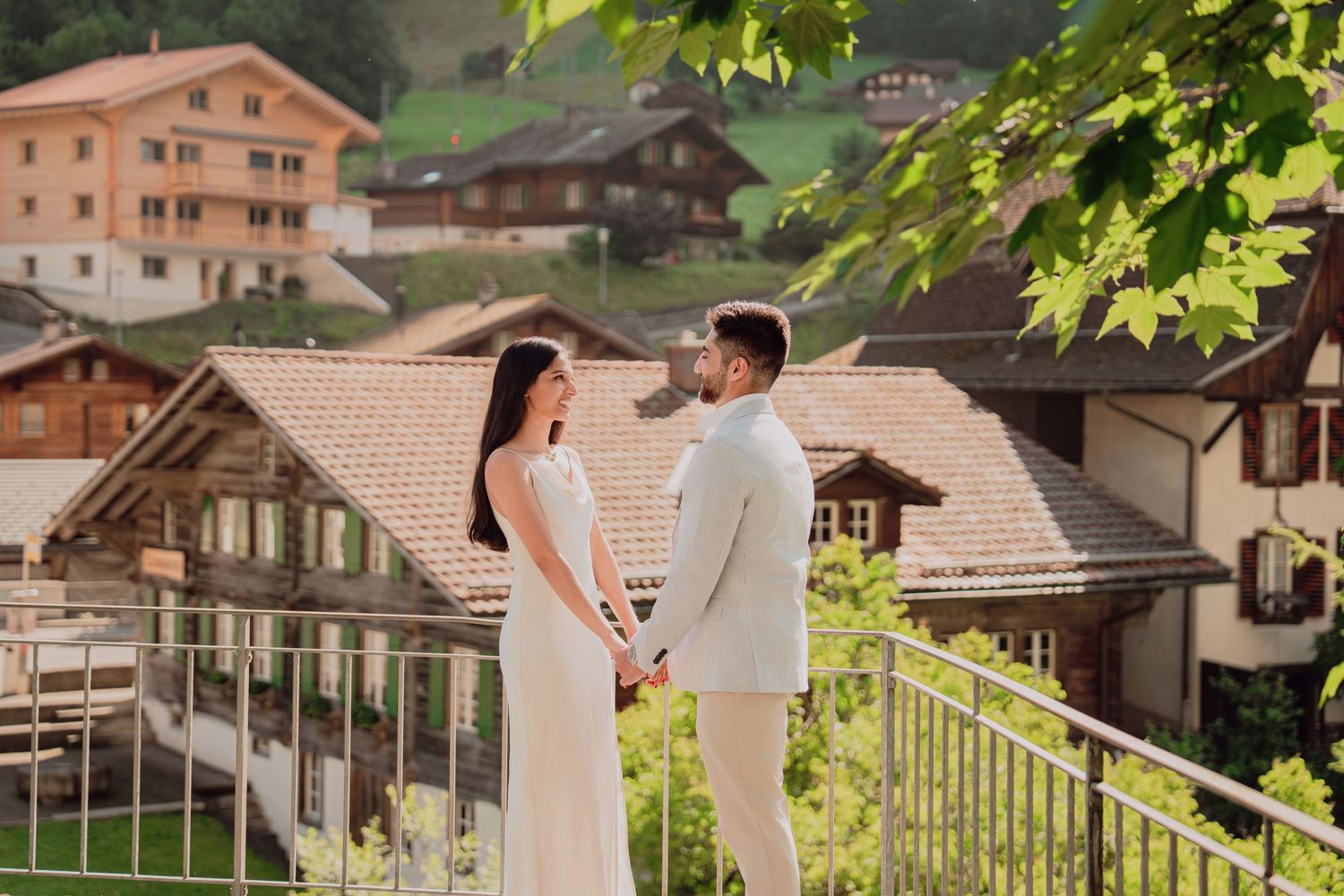Bride and groom hold hands on a balcony overlooking traditional Swiss chalet-style homes in a mountain village.