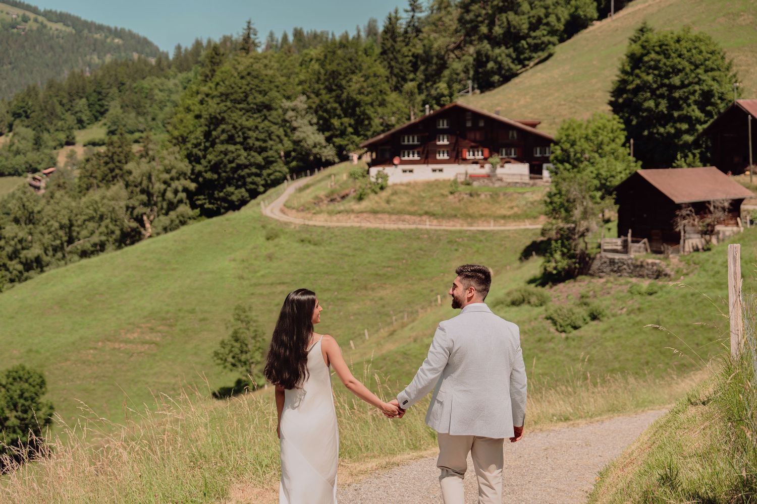 Bride and groom holding hands walking toward a traditional Swiss chalet on a green hillside path.