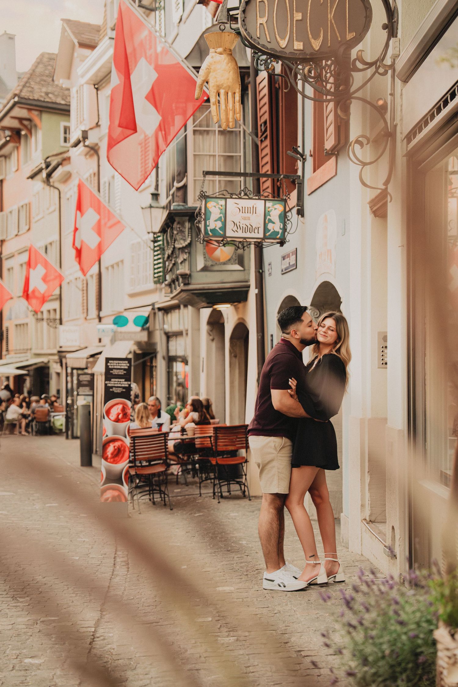 Couple sharing a kiss on a picturesque Swiss street with red flags overhead.