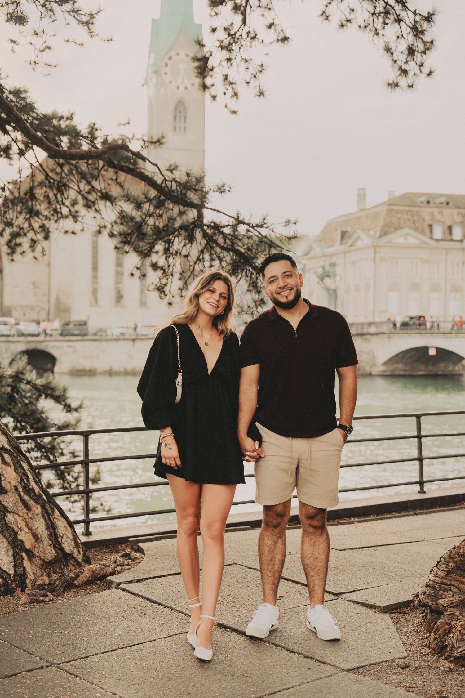 Couple standing together by Zurich river with Fraumunster church tower visible in background.