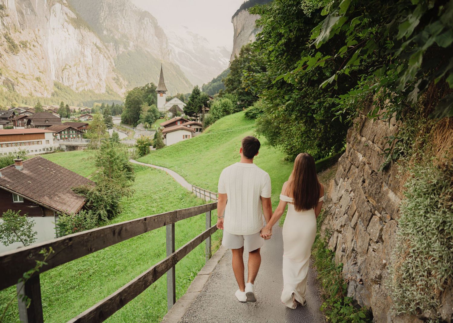 Couple holding hands walking down a scenic path with a stunning Swiss alpine village and green meadows in background.