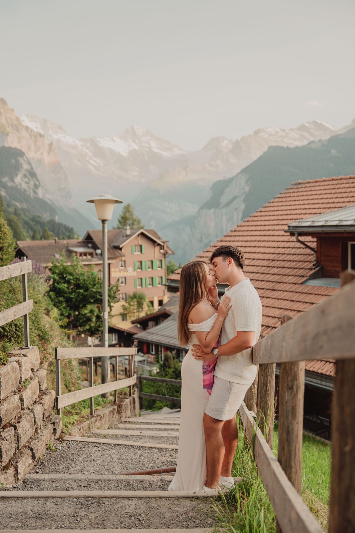 Couple kissing on stone staircase in picturesque Swiss Alpine village with snow-capped mountain peaks behind.