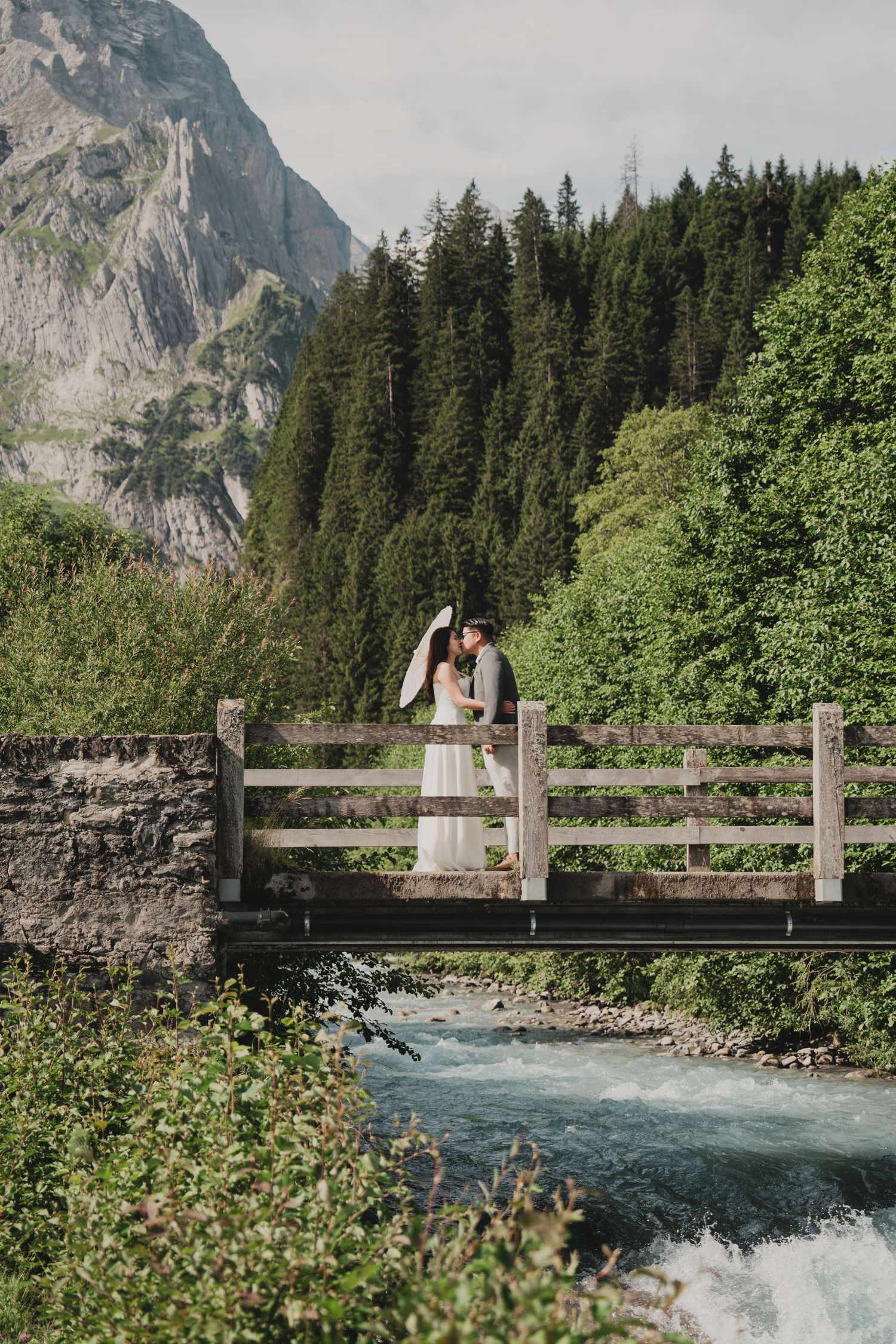 Wedding couple embraces on wooden bridge over glacial river with dramatic mountain backdrop and forest.