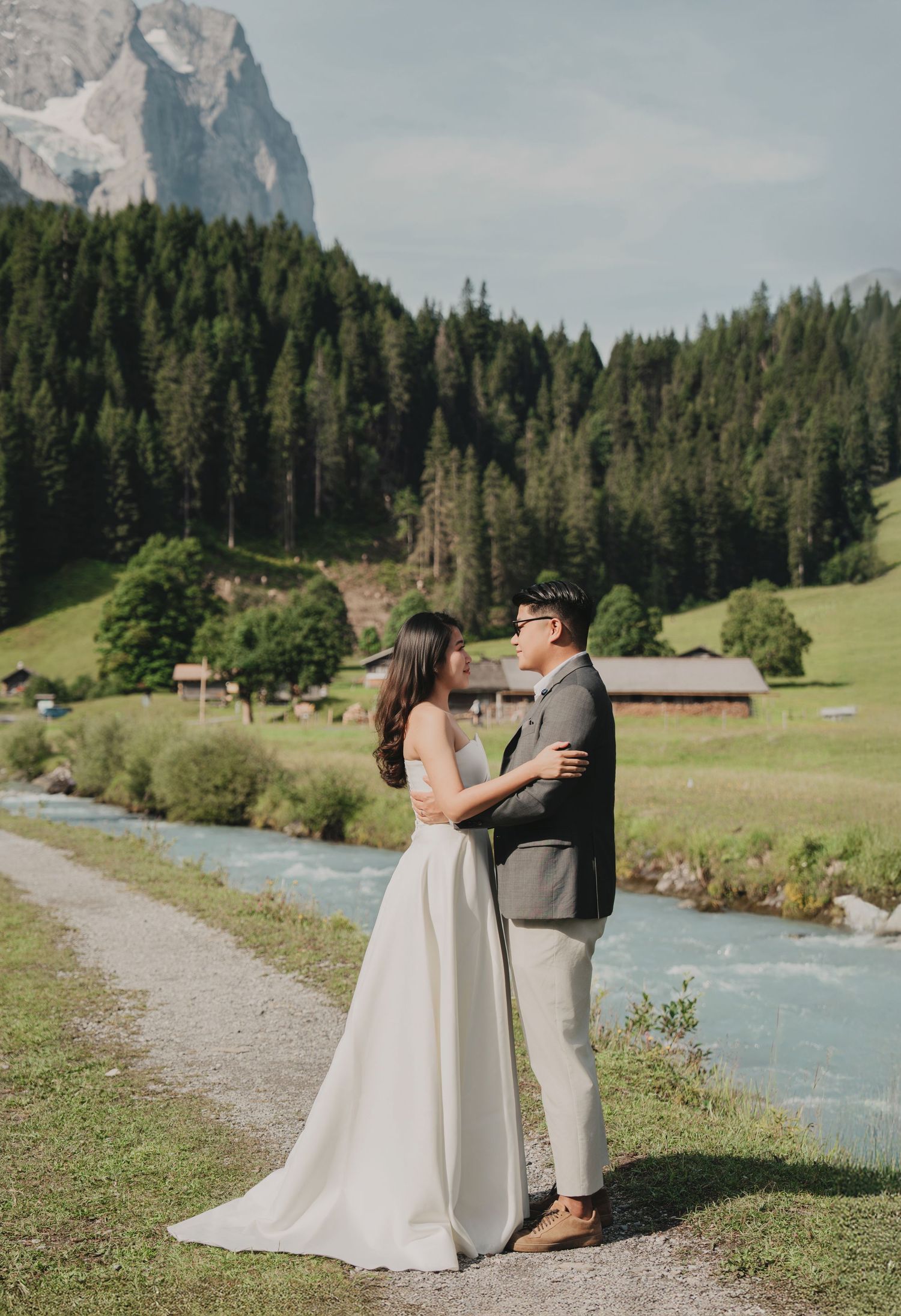 Wedding couple holding each other beside a rushing alpine river with pine forests and mountain peaks behind them.