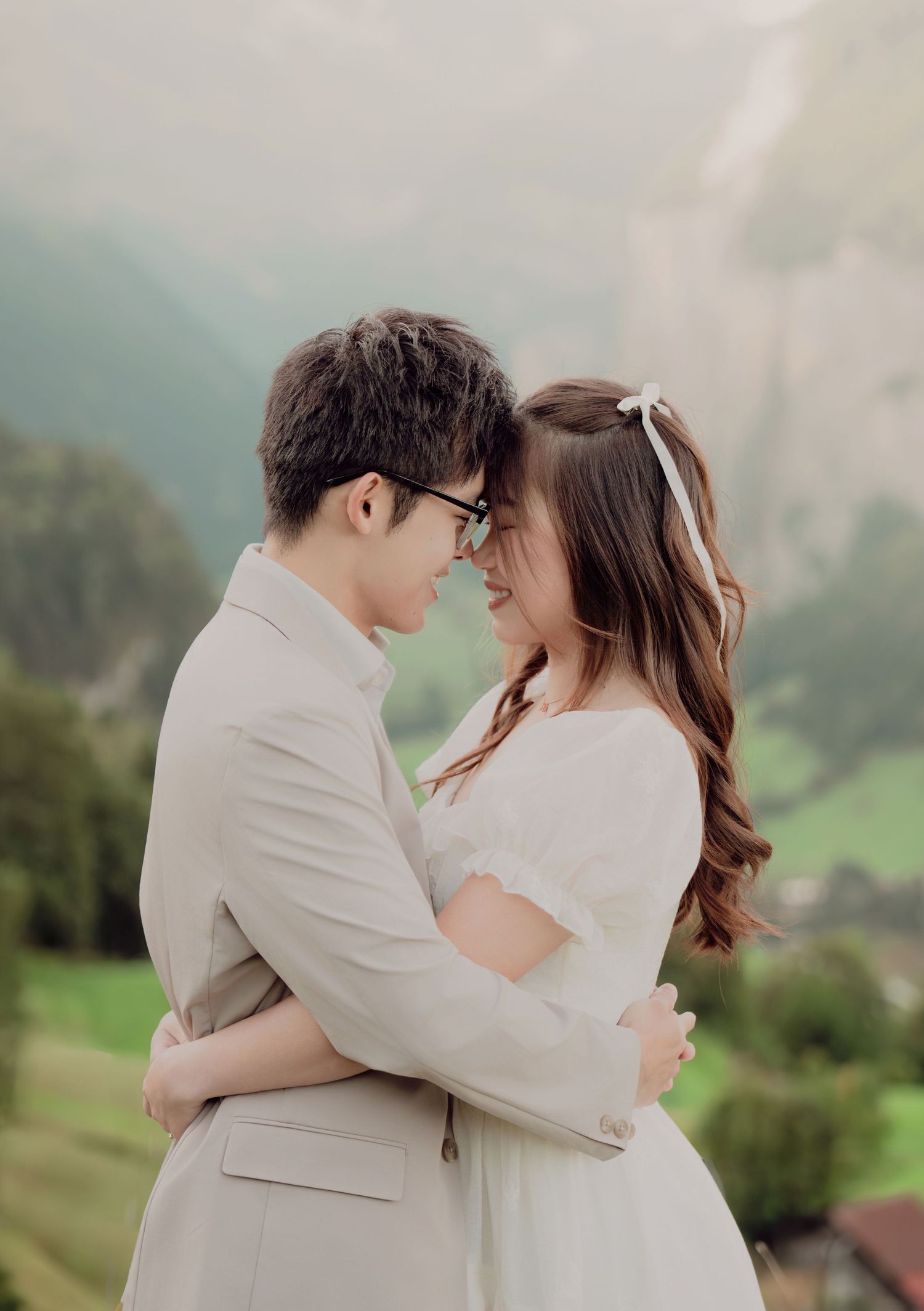 Couple embracing intimately in white dress and beige suit with misty green Swiss Alps valley backdrop.