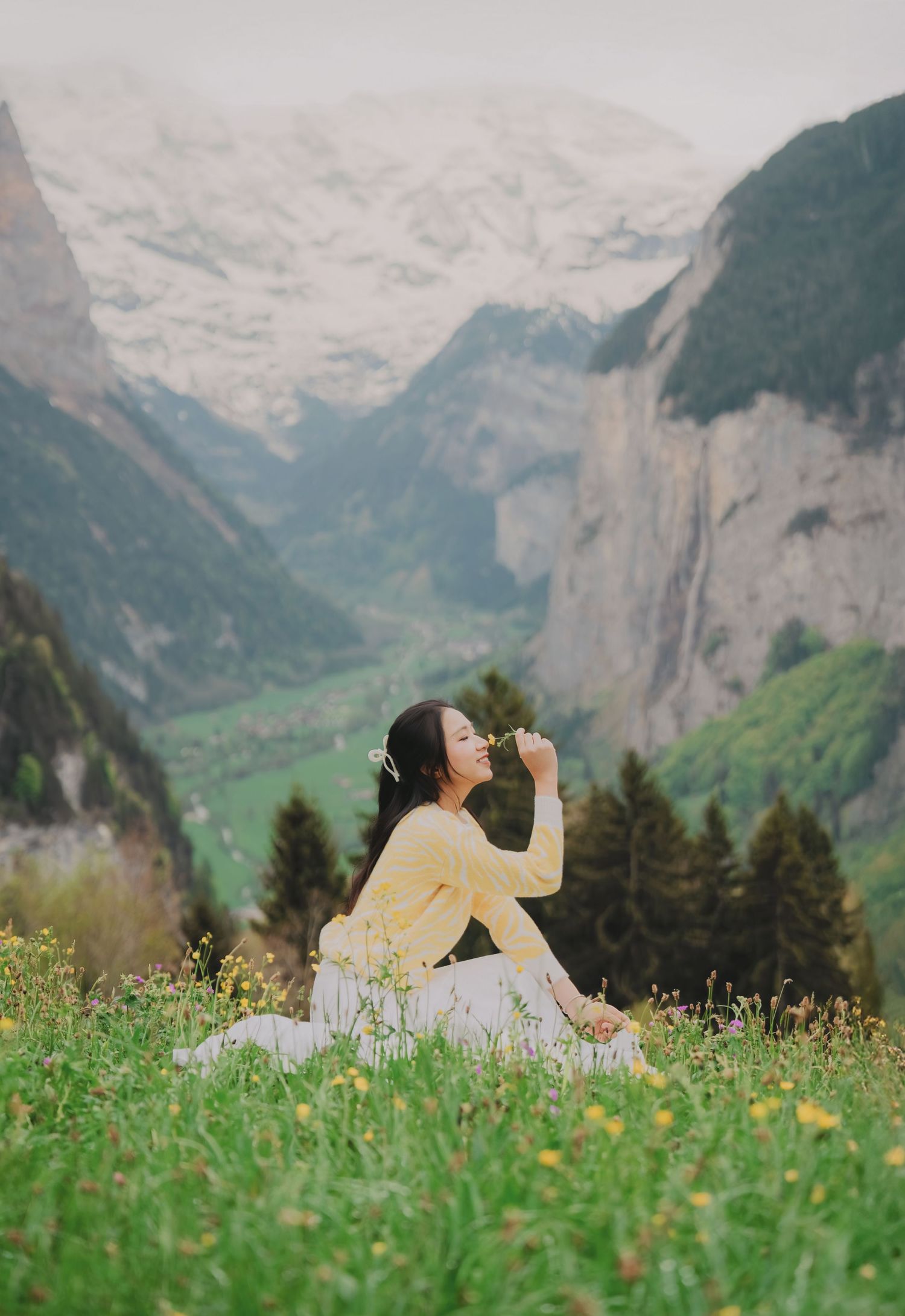 Young woman in yellow cardigan sitting in alpine meadow smelling flower with snowy mountains behind.