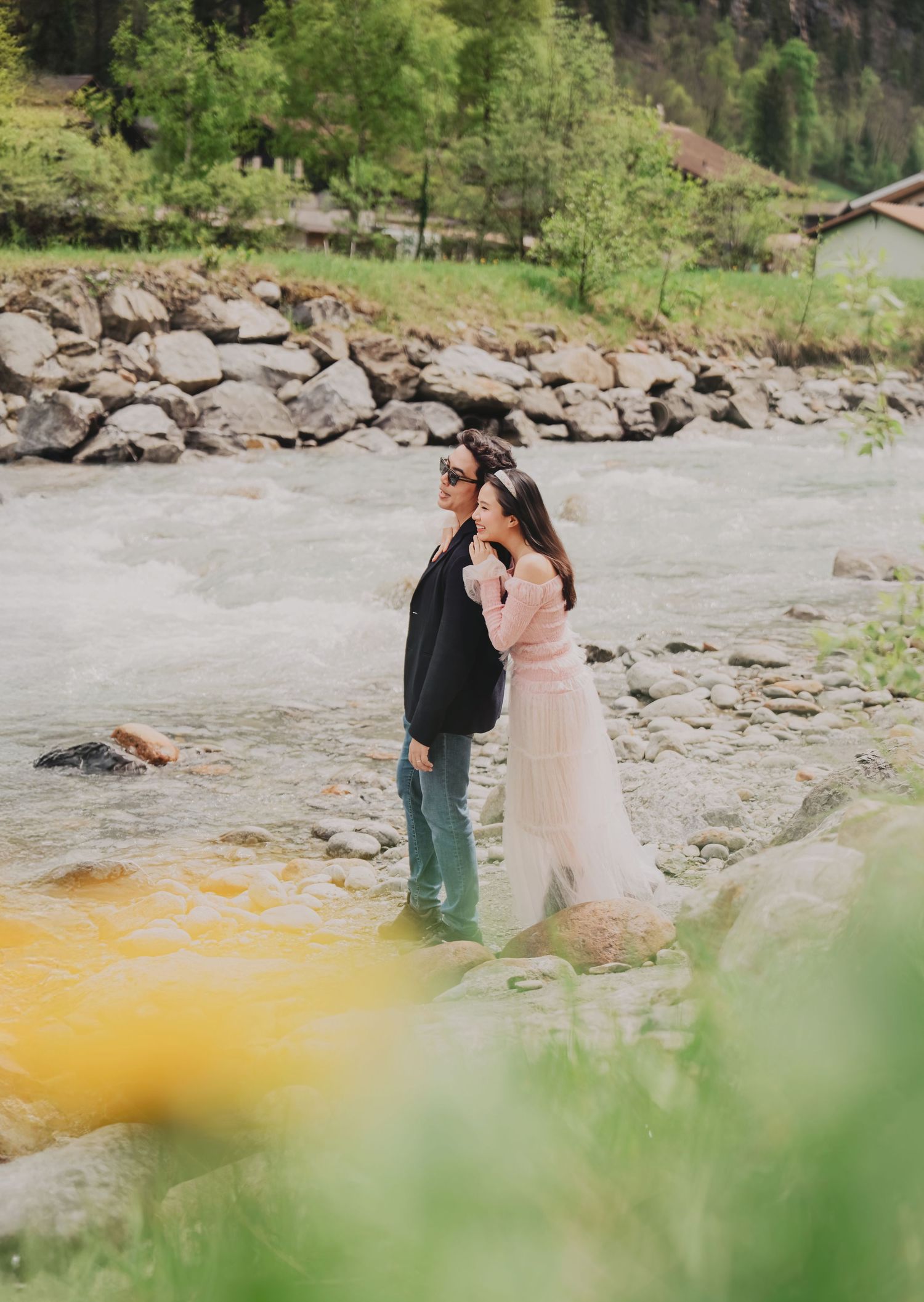 Couple embracing romantically on rocky riverbank with rushing alpine stream and stone wall backdrop.