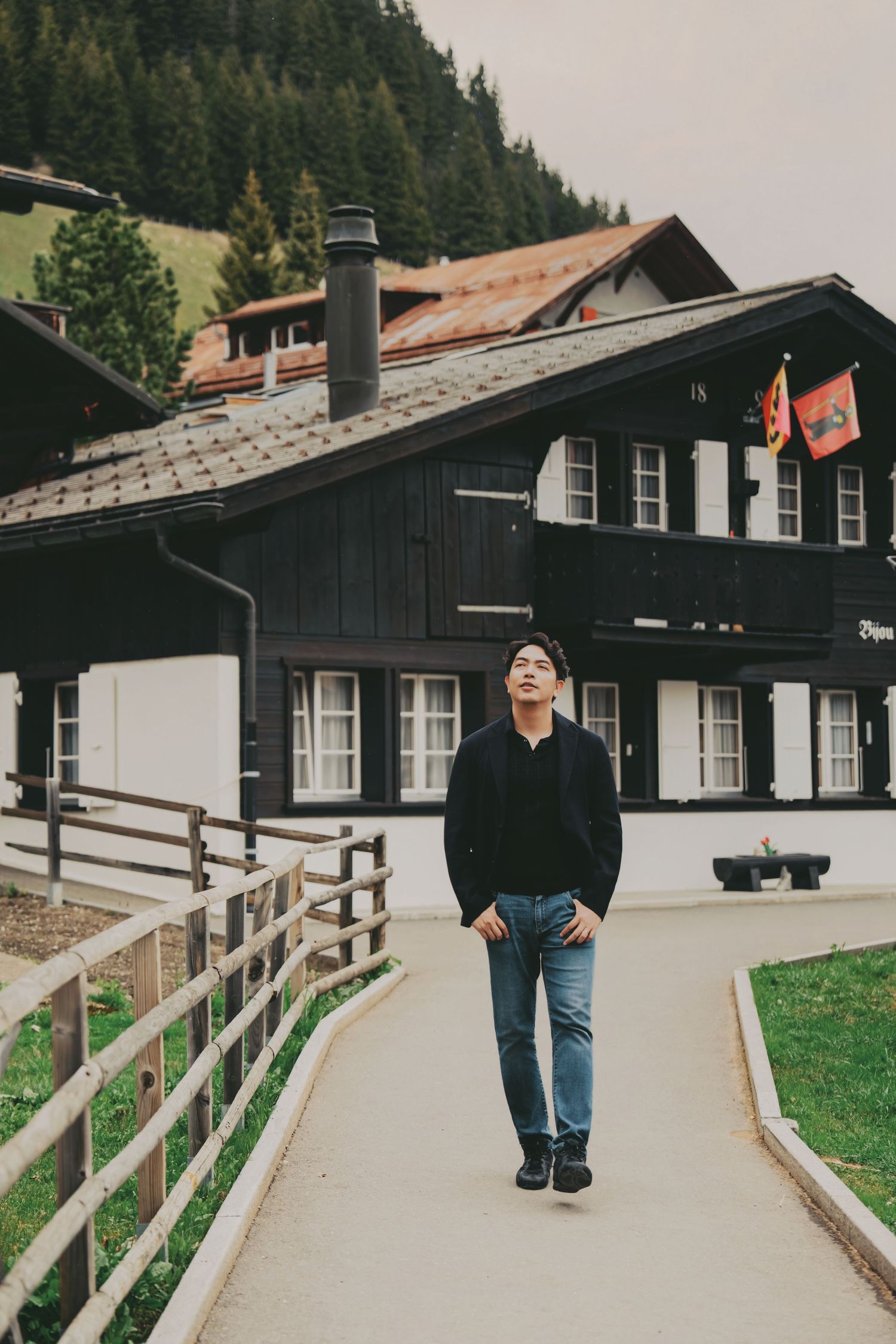 Young Asian man in black sweater walking on path before traditional dark Swiss chalet with forest backdrop.