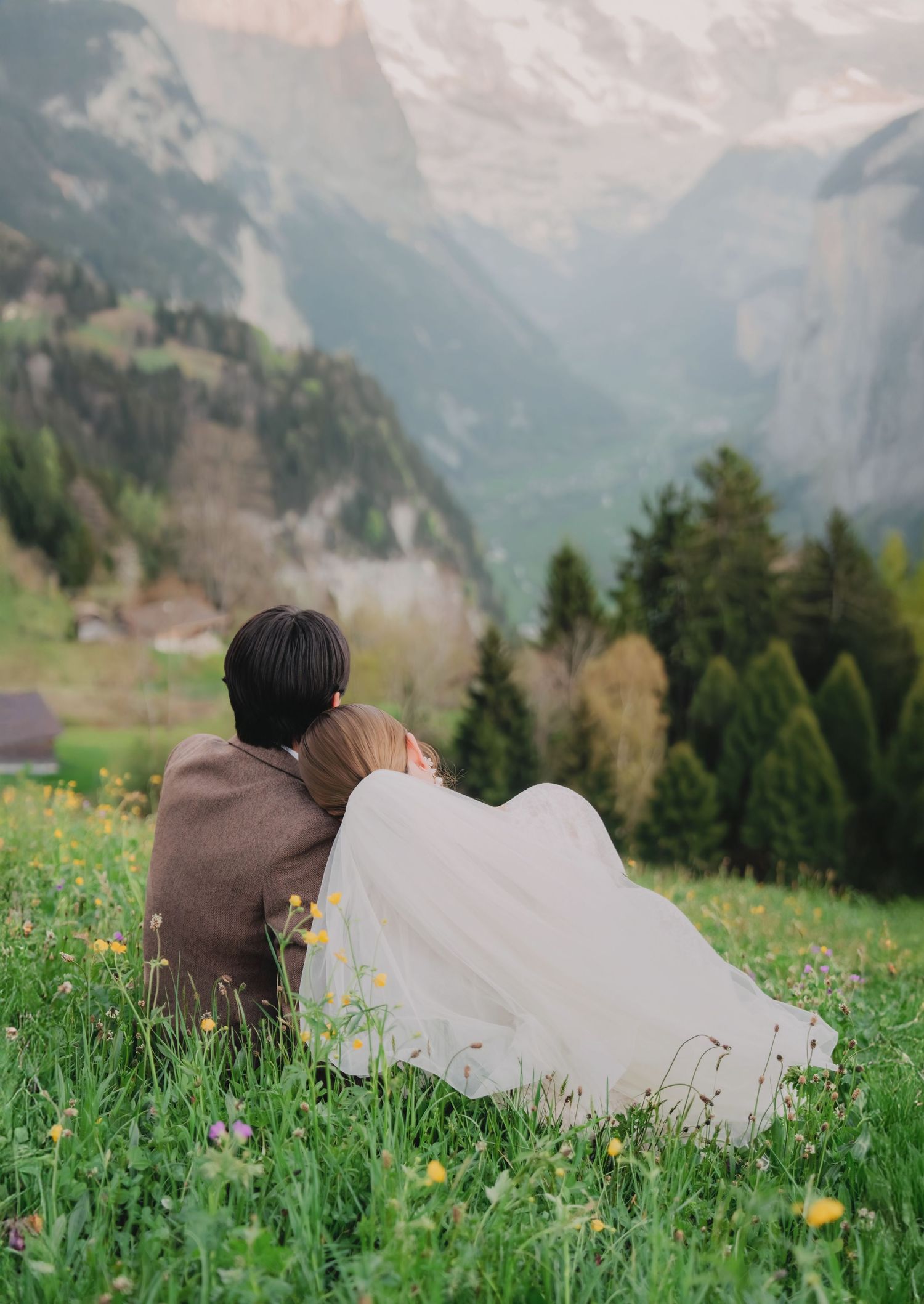 Newlyweds sitting together on flower meadow admiring breathtaking Swiss mountain valley scenery.