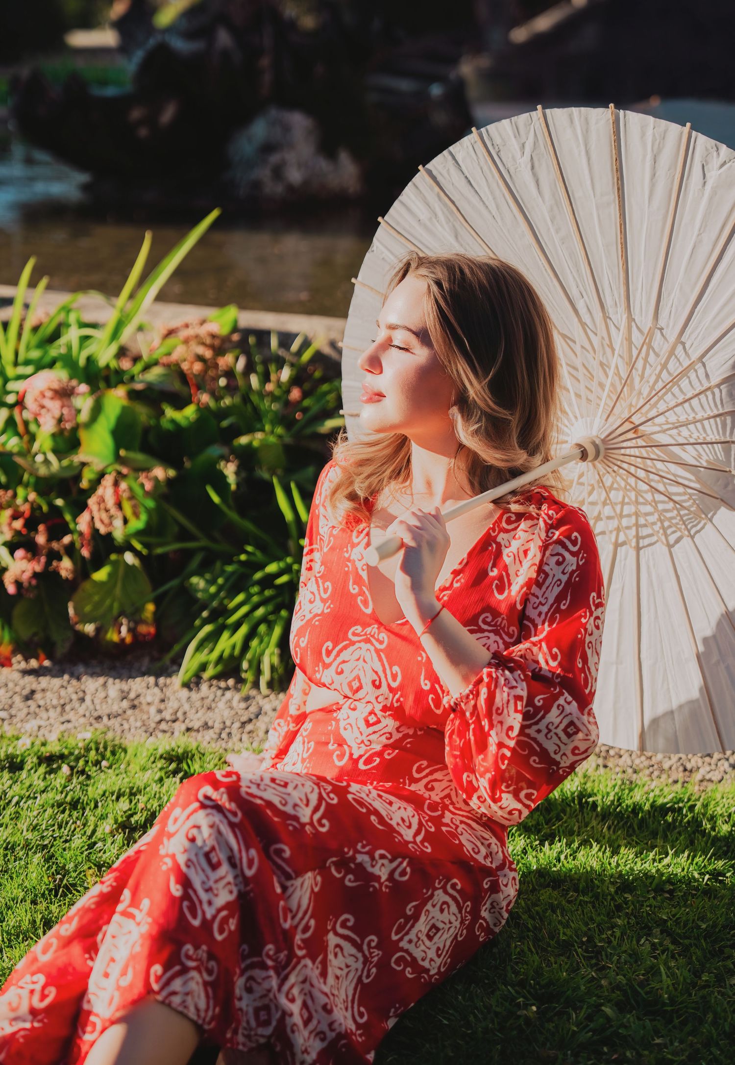 Woman in red patterned dress sitting on grass with white parasol.