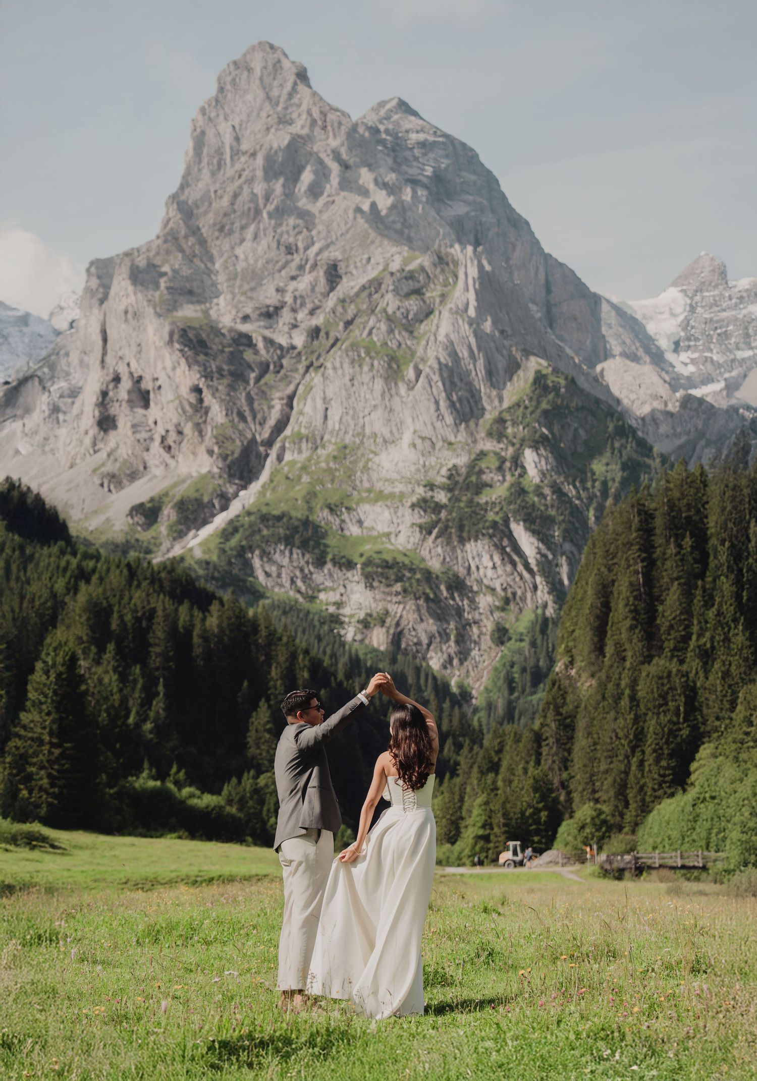 Groom spinning bride in white dress on green meadow with stunning Swiss Alps mountain scenery.