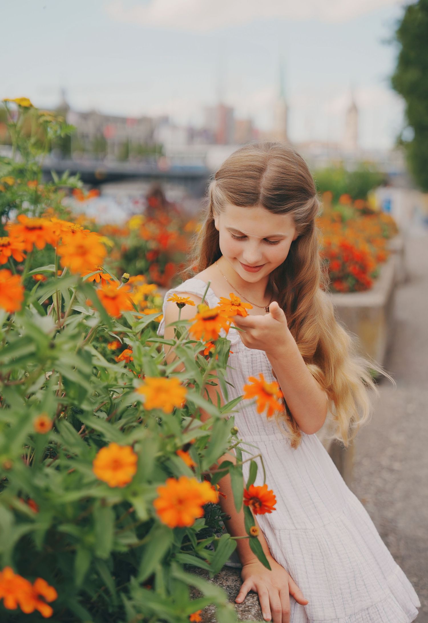 Young blonde girl in white dress smelling orange marigold flowers along a European city promenade.