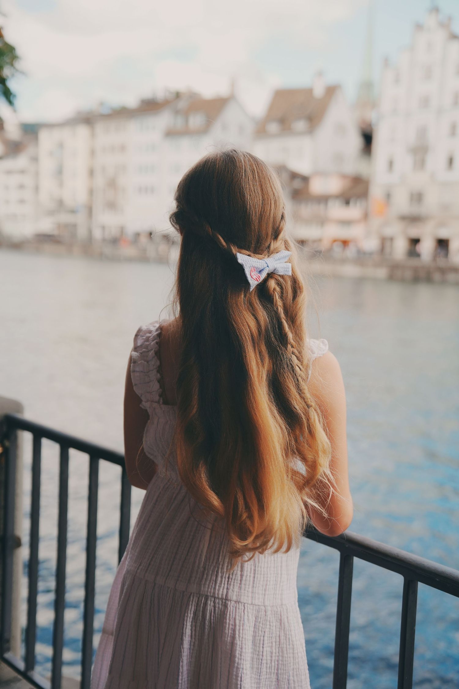 Girl with long hair and bow clip in pink dress stands at railing overlooking European river city.