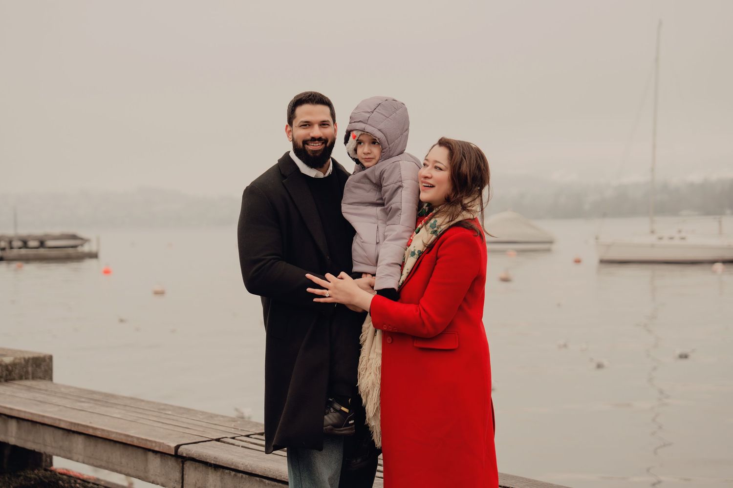 Happy family of three posing by a foggy lake, father holding child, mother in red coat smiling.