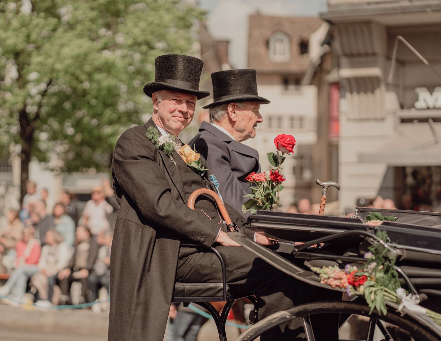 Two elegantly dressed men in top hats ride in a classic black carriage at a parade.