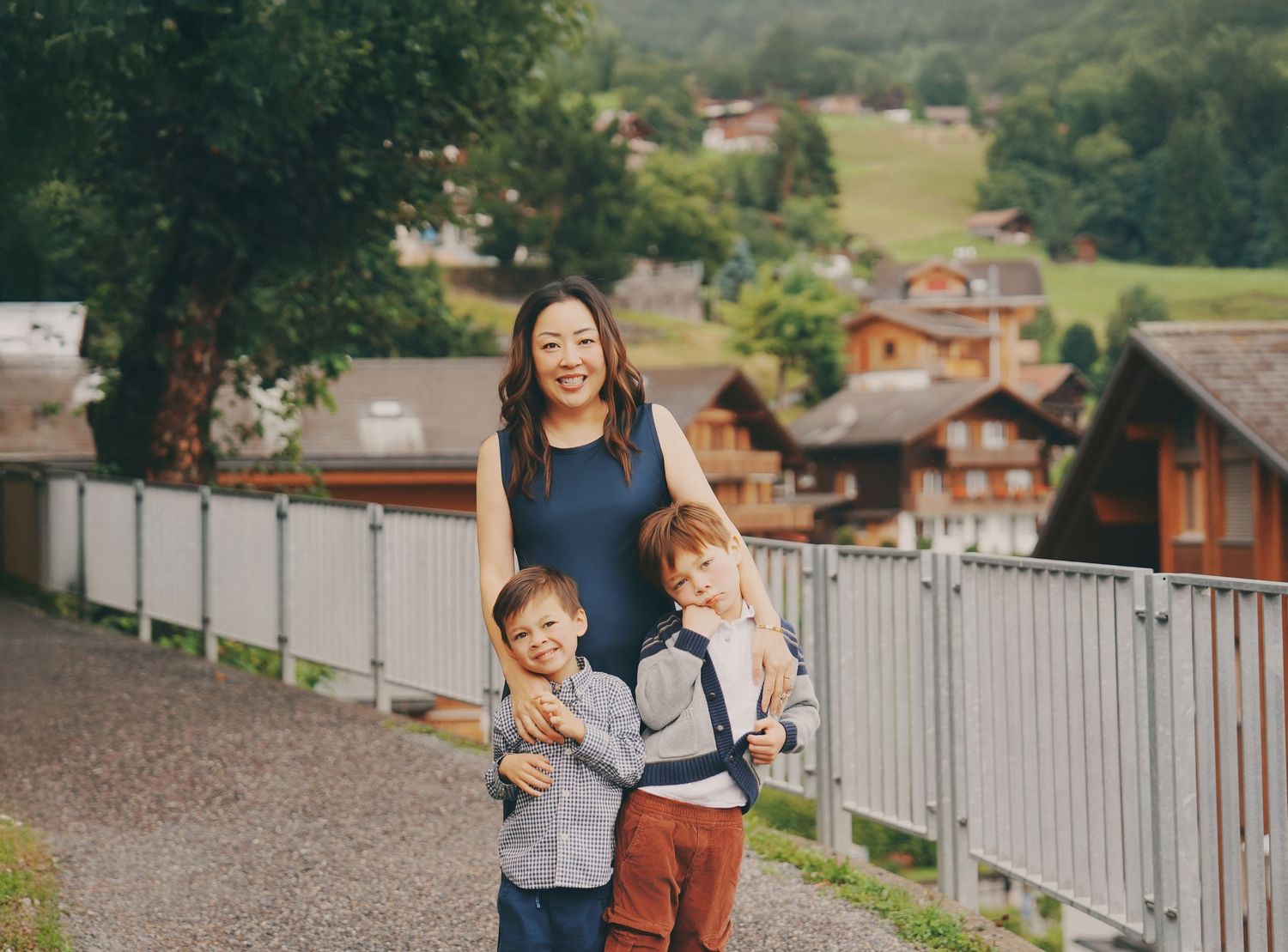A smiling Asian woman poses warmly with two young boys on a Swiss village pathway.