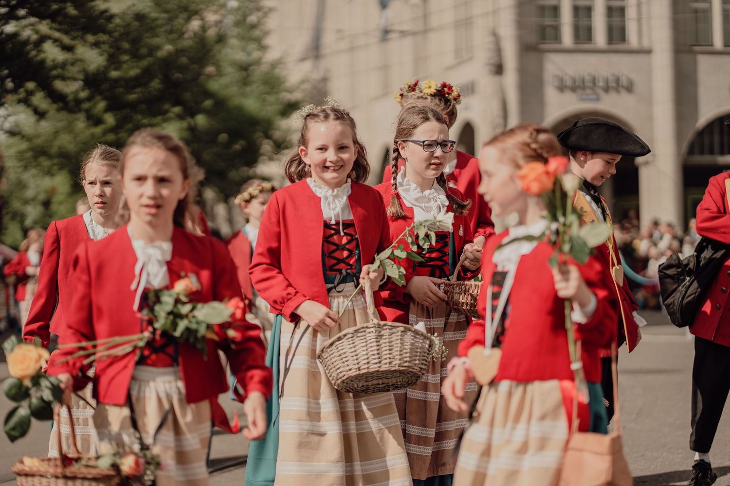 Girls in red and white traditional folk costumes carry flower baskets during a festive street parade.