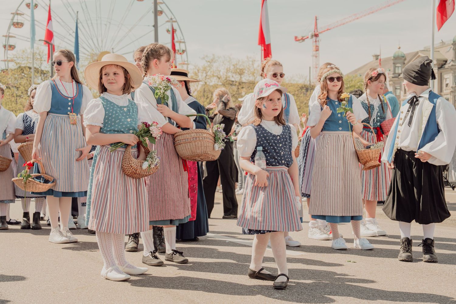 Girls in traditional Swiss dirndl dresses carrying wicker baskets walk in a sunny outdoor parade.