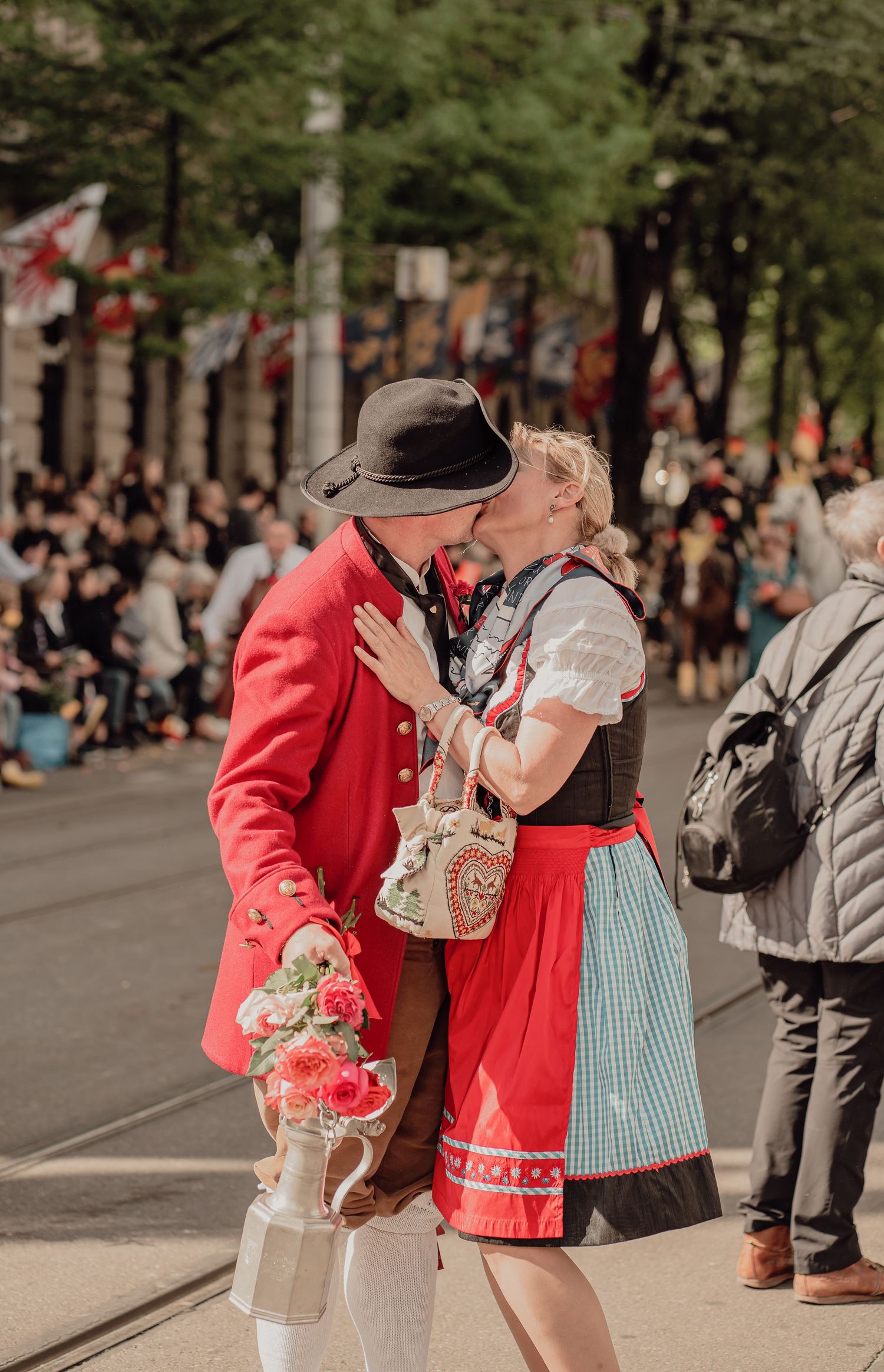 Man in red historical jacket kisses woman in dirndl dress during a traditional street festival.