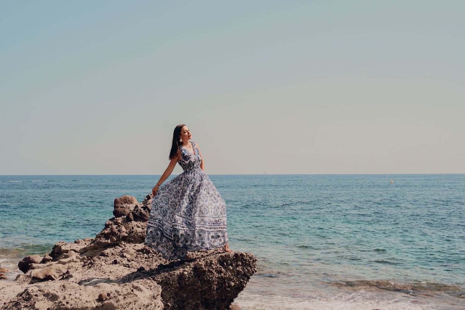 Woman in blue floral maxi dress sitting on ocean rocks under a clear sky.