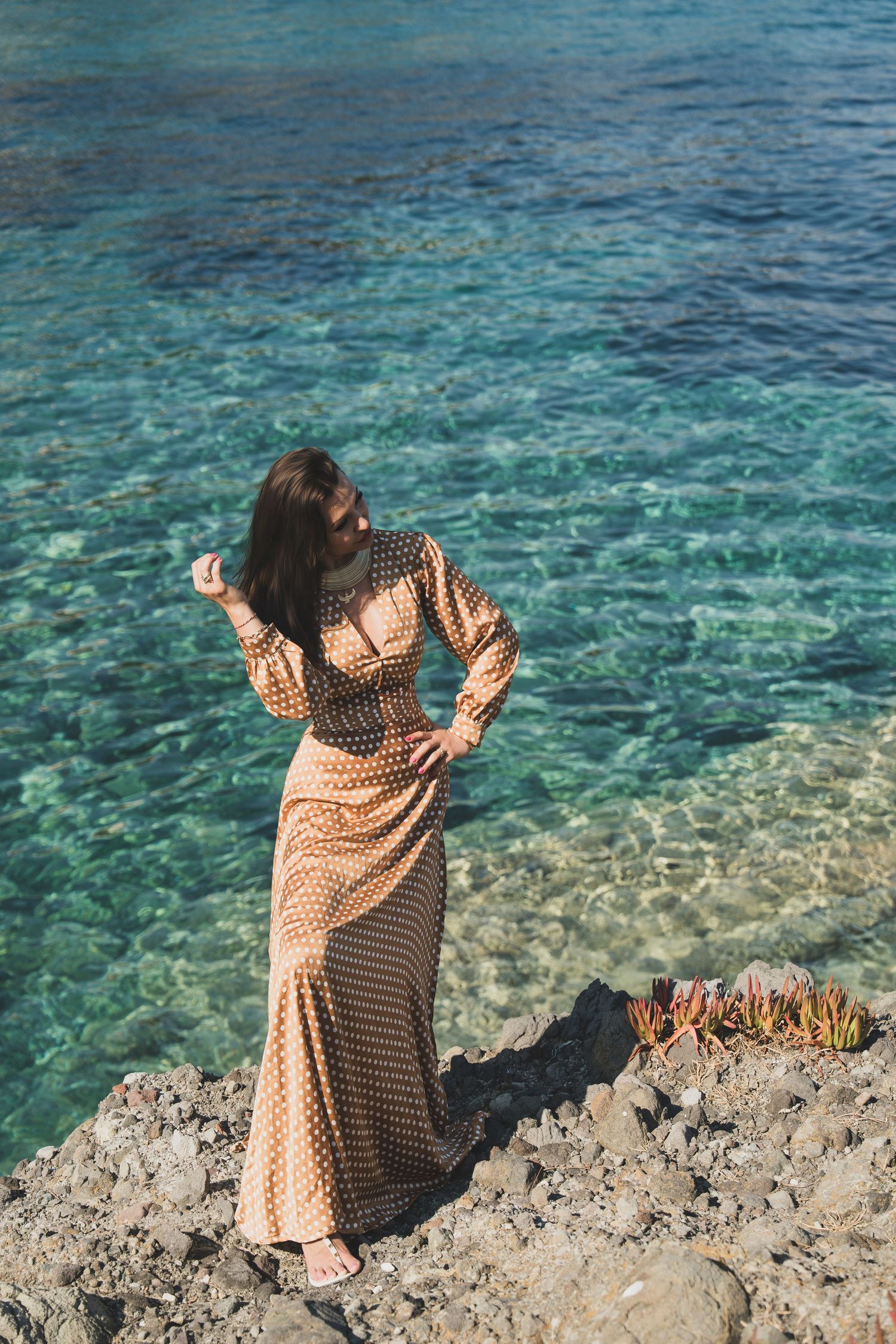 Brunette model in gold polka dot dress posing by crystal clear turquoise ocean water.