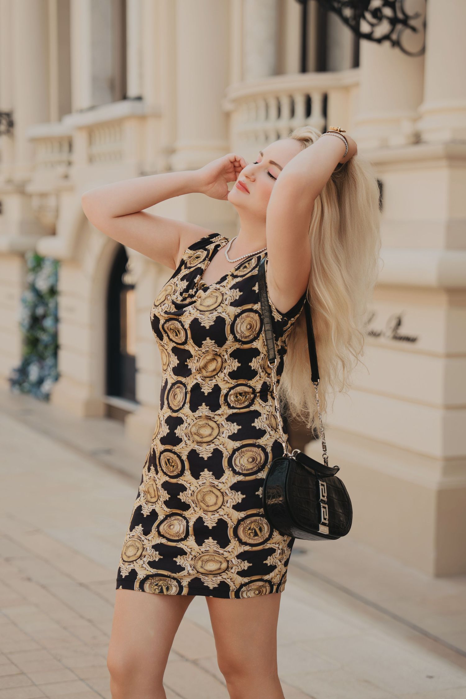 Blonde woman in black and gold Versace print mini dress posing on elegant European street.