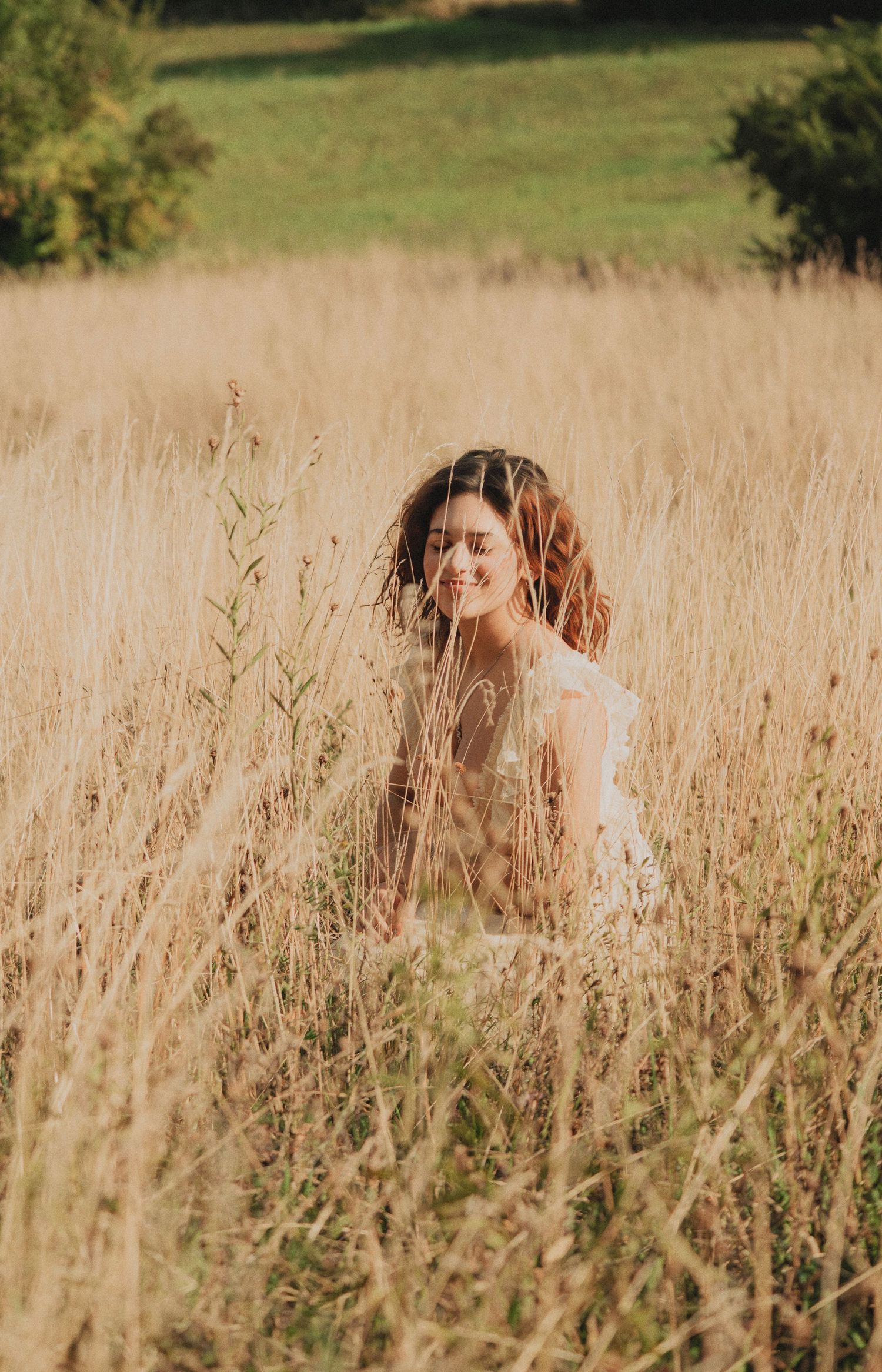 Smiling woman in white dress sitting among tall golden dry grass in sunny field.