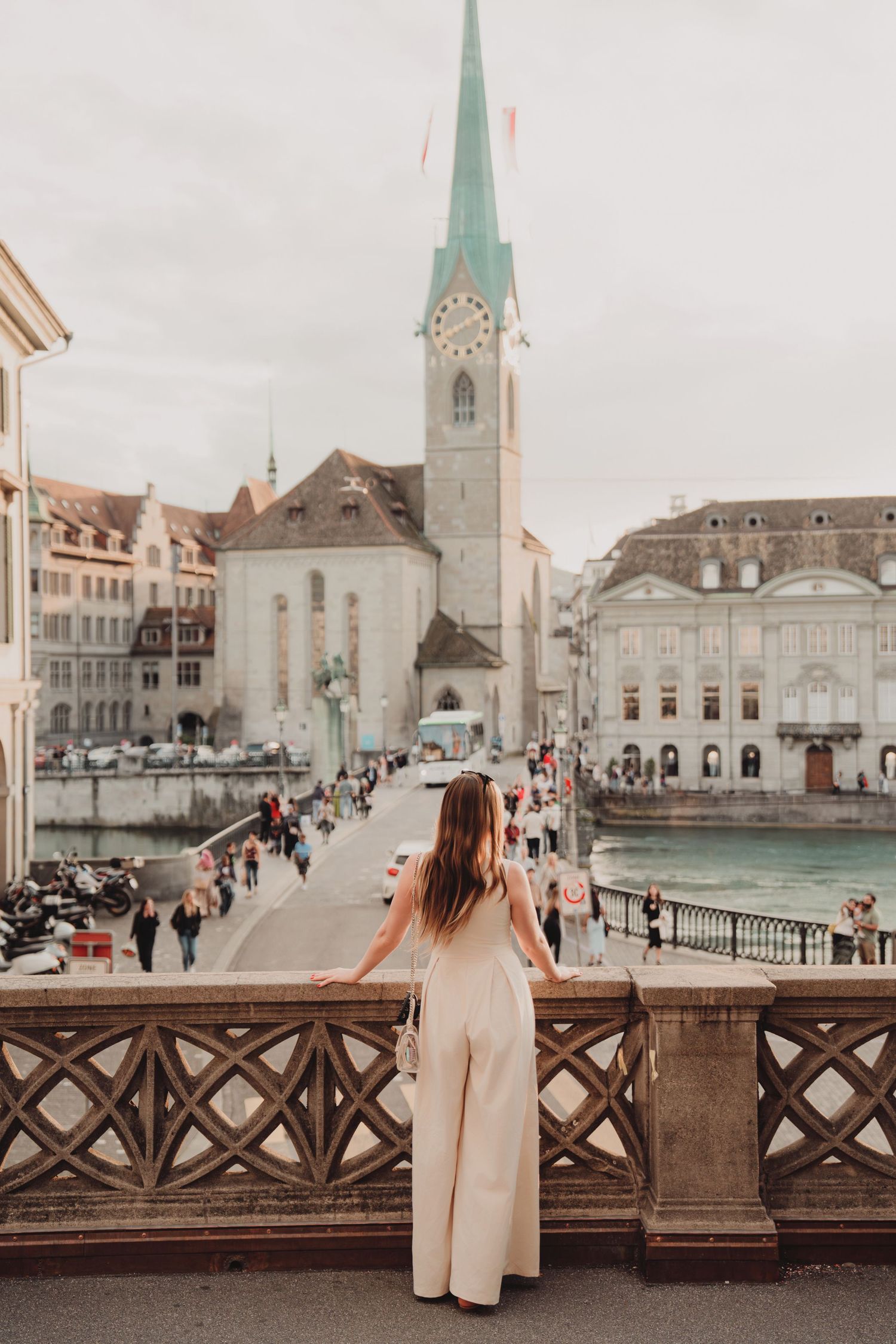 Woman in cream jumpsuit stands on ornate bridge overlooking Zurich's Fraumunster Church and Limmat River.