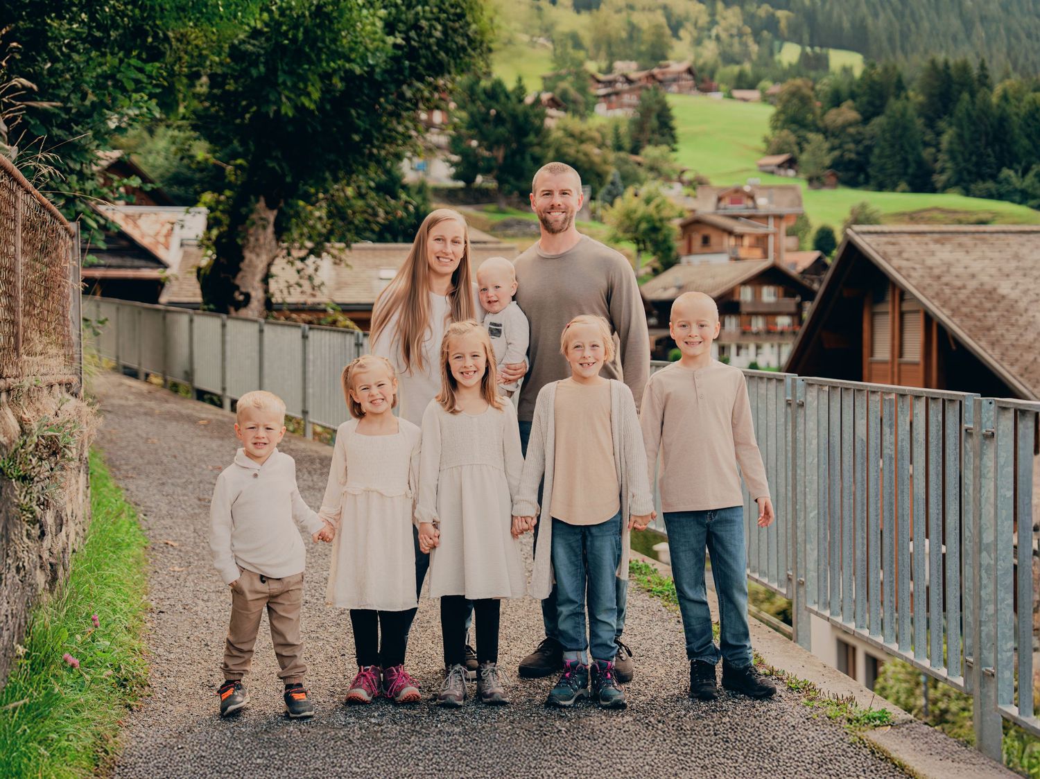 A happy family of eight poses together on a scenic gravel path in a Swiss mountain village.