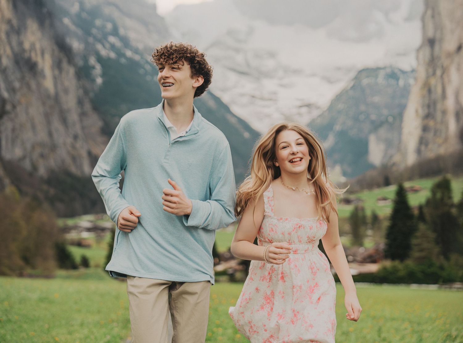 Teenage couple laughing and running together in a green Alpine meadow with dramatic mountain cliffs behind them.