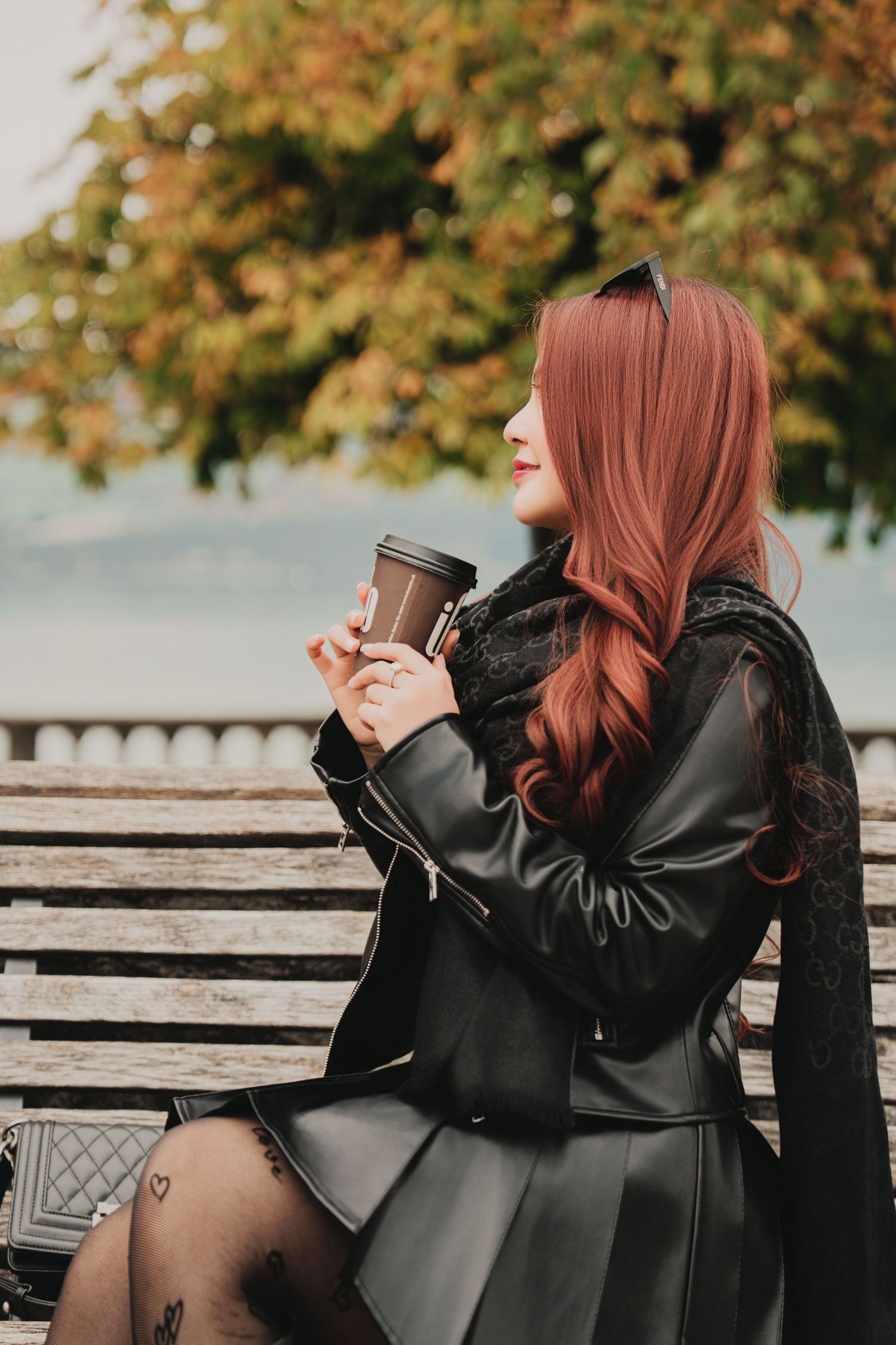 Smiling red-haired woman in black leather jacket and mini skirt sits on bench holding takeaway coffee cup outdoors.