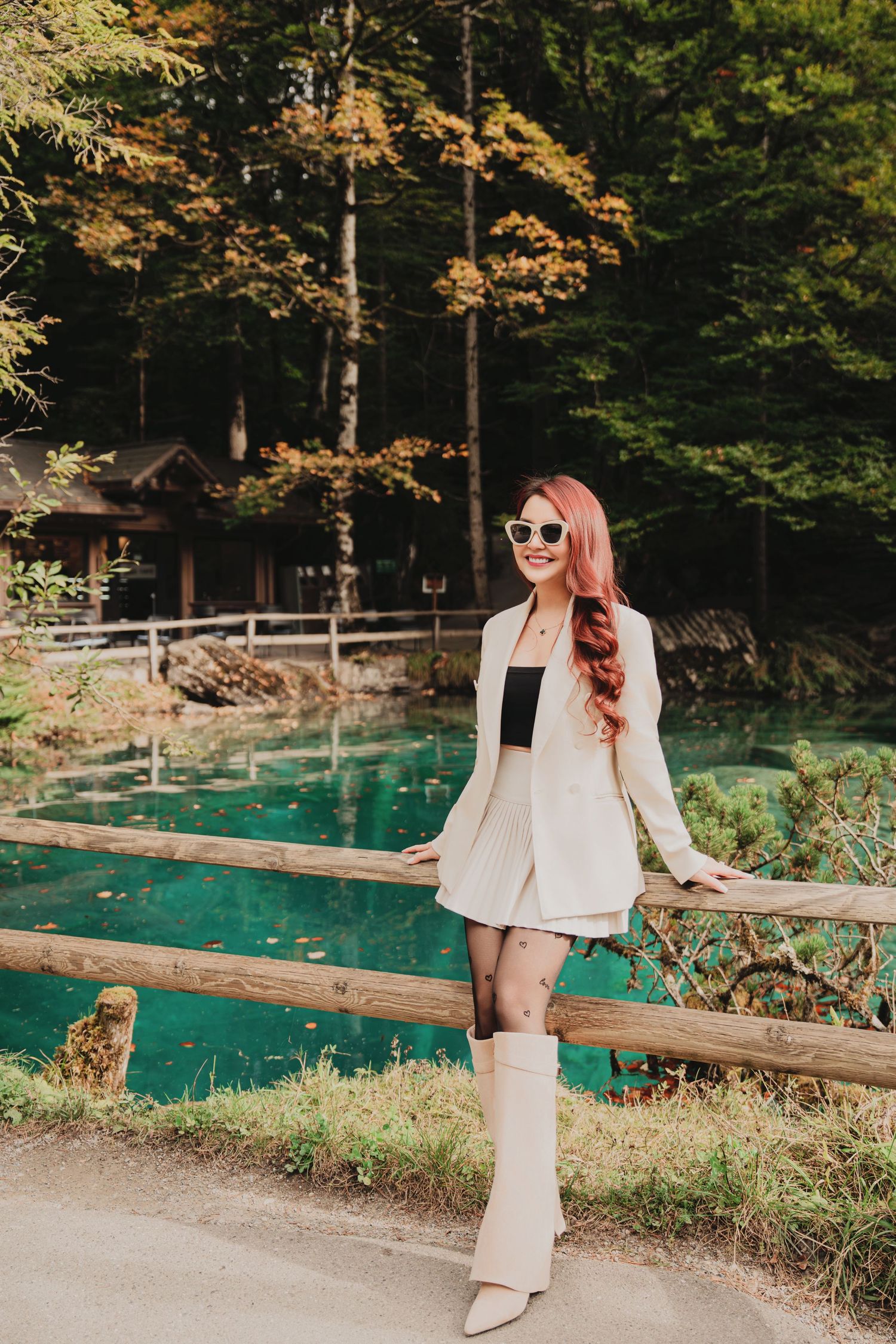 Stylish woman in white suit and boots posing by emerald green lake in fall forest.