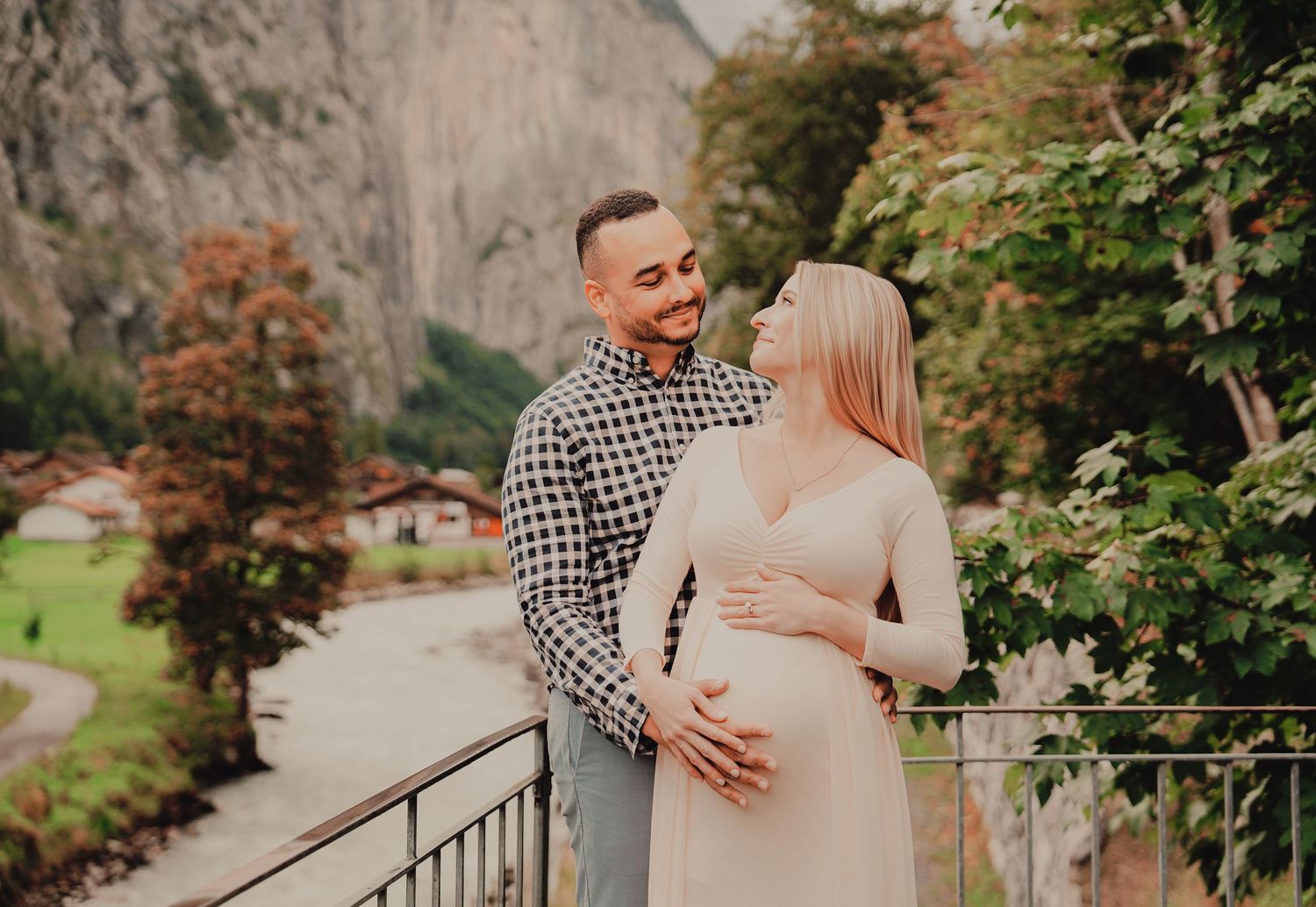 Couple sharing tender moment on bridge, pregnant woman in beige dress with rocky cliffs behind them.