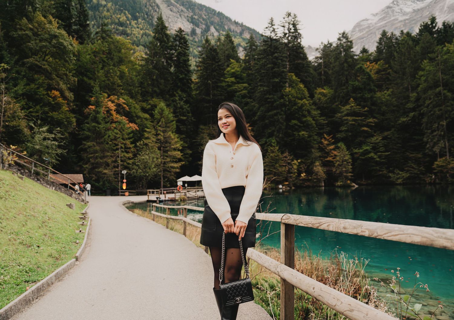 Smiling woman in casual white sweater and black pants walks along turquoise lakeside path.