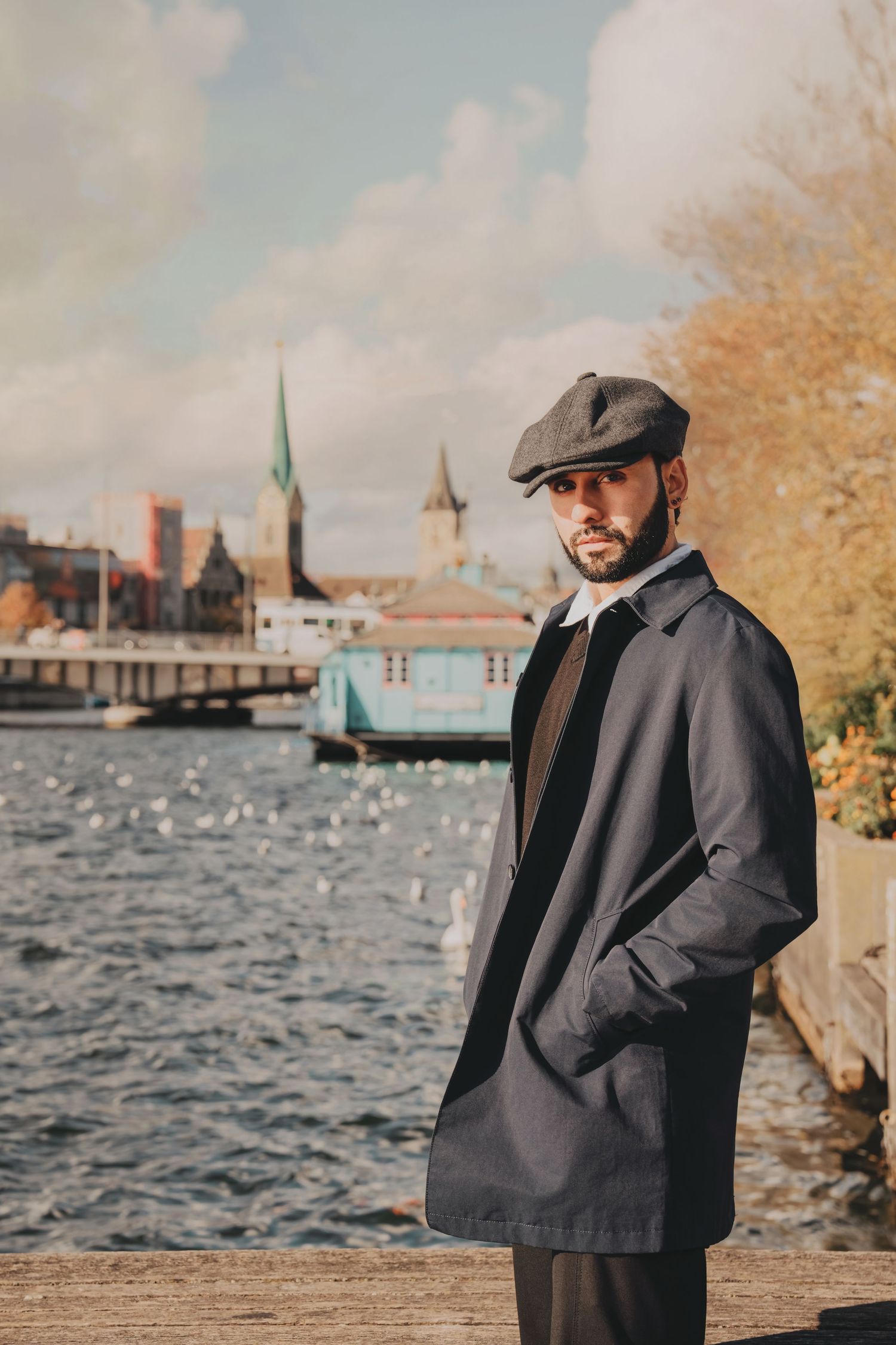 Stylish bearded man in navy trench coat and flat cap stands by river with historic city backdrop.