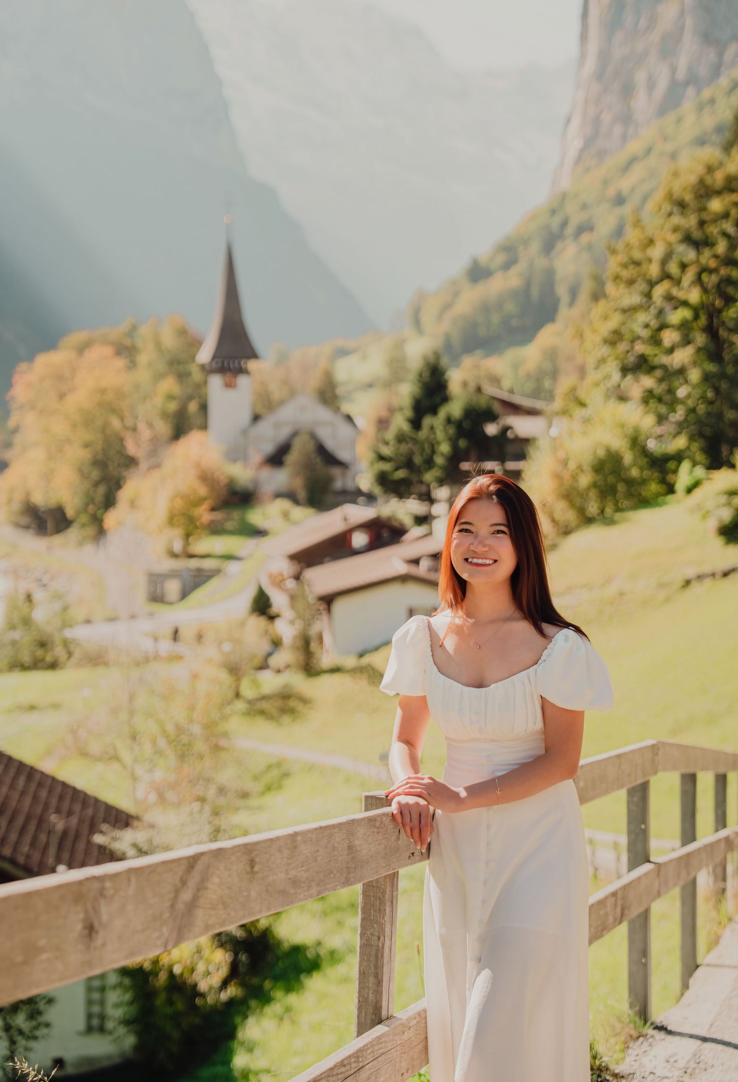 A smiling woman in a white dress stands on a balcony with a beautiful Swiss alpine village behind her.