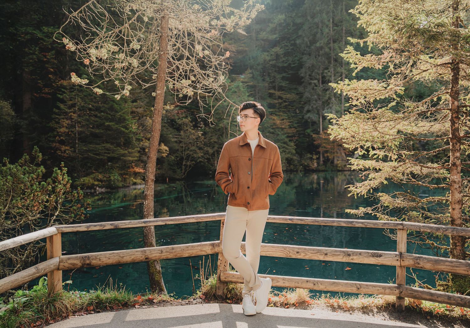 Young man in a brown jacket standing on a wooden deck overlooking a stunning turquoise forest lake.