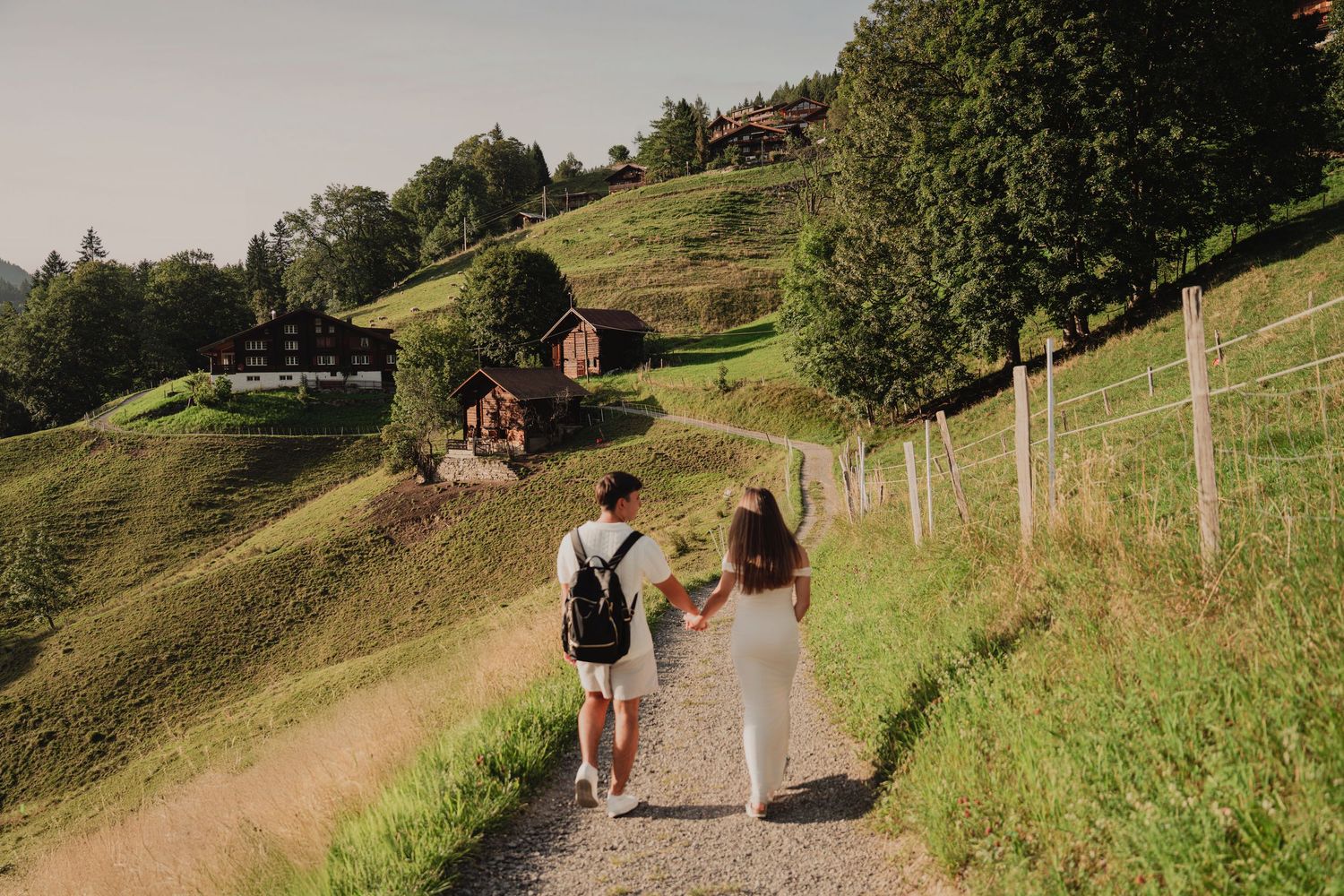 A couple walks together on a scenic mountain path carrying flowers in a green alpine valley.