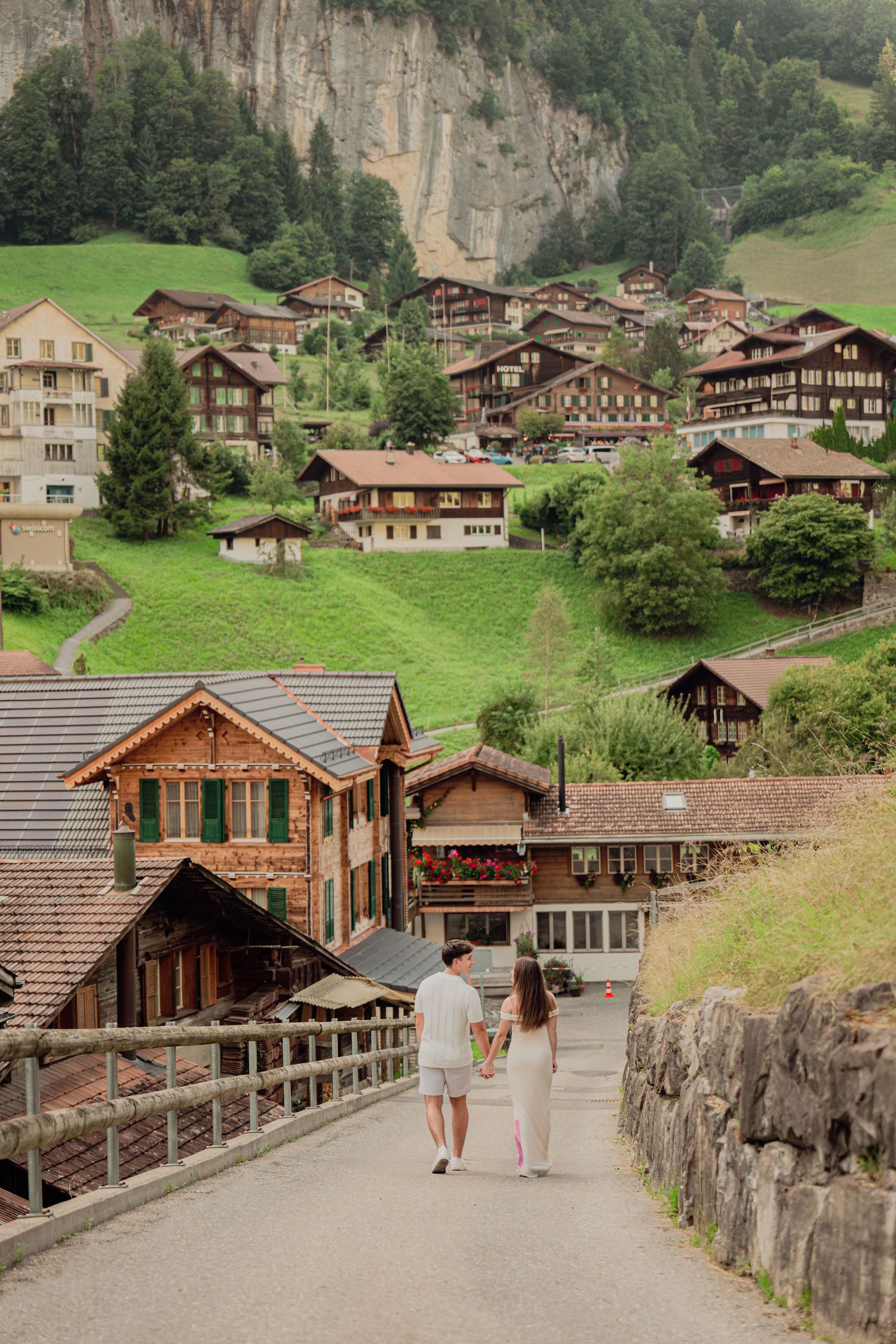 Couple walking hand in hand through charming Swiss alpine village with traditional wooden chalets on green hillside.