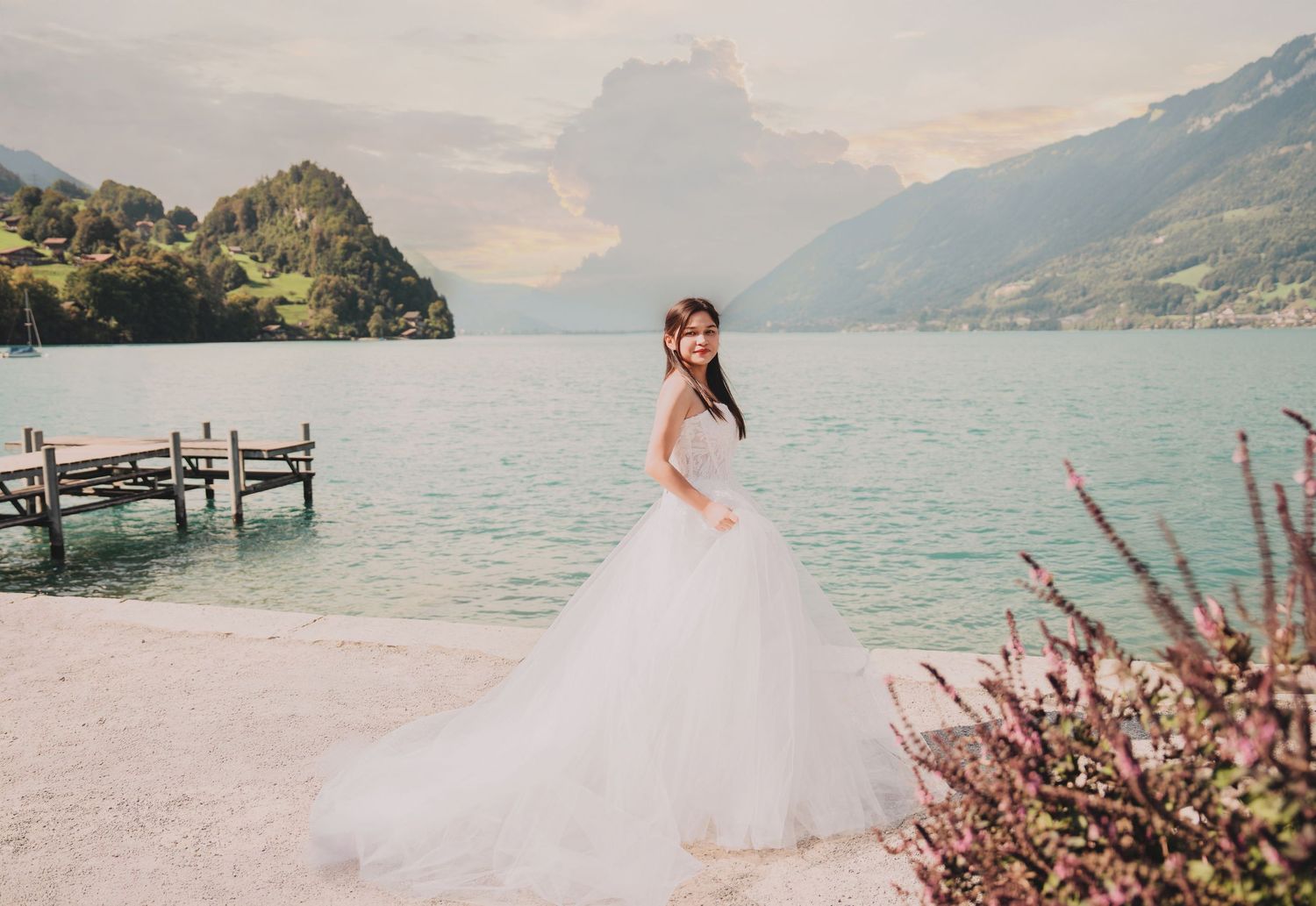 Elegant bride in full tulle wedding gown stands on lakeside with alpine mountains and wooden dock behind her.