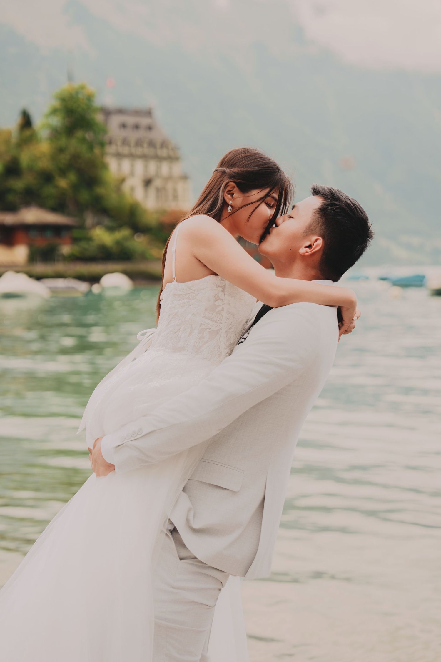 Groom lifting and kissing bride by a picturesque alpine lake with boats and a grand building in background.