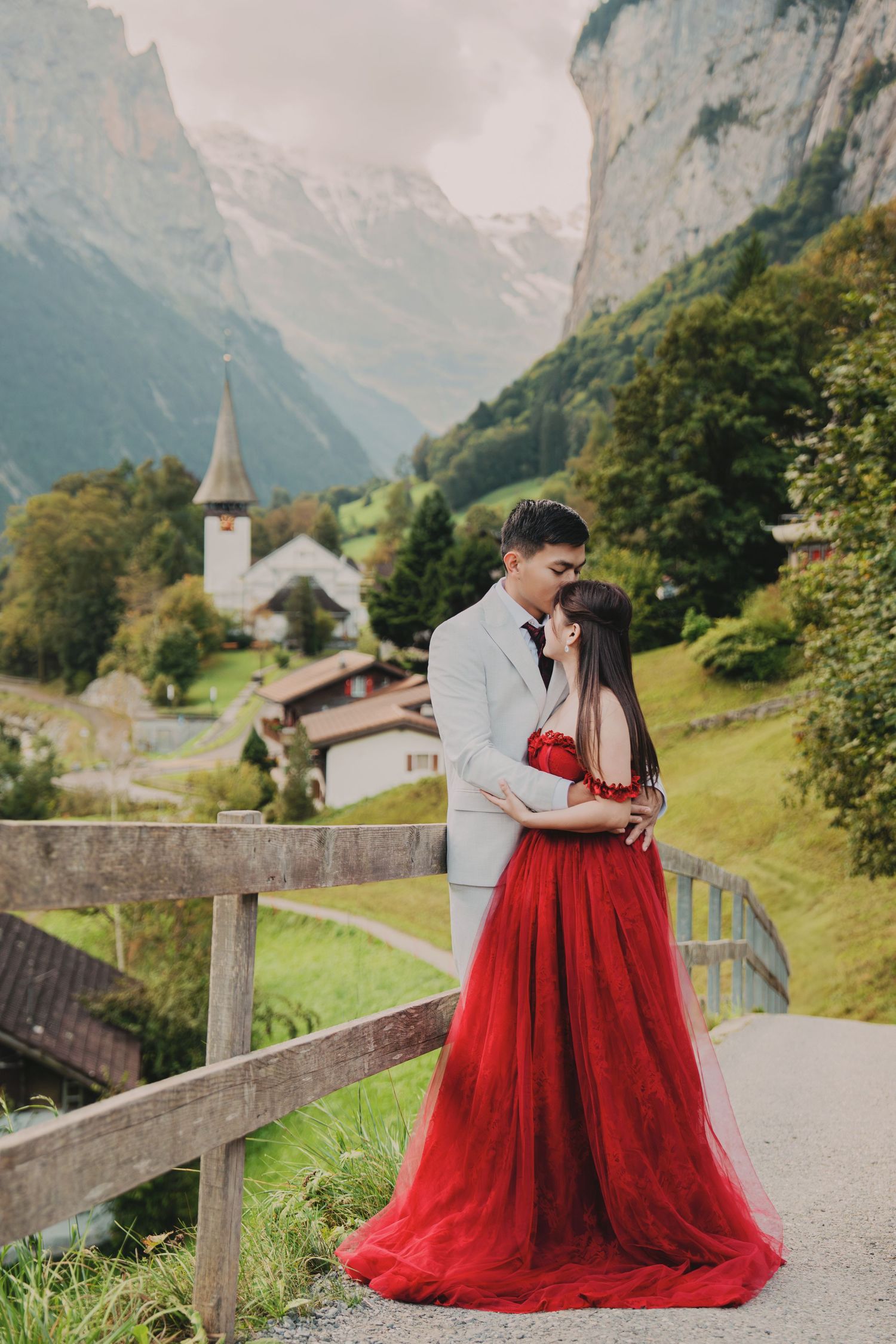 Couple sharing a romantic moment in red tulle gown and grey suit with Swiss Alps village scenery.