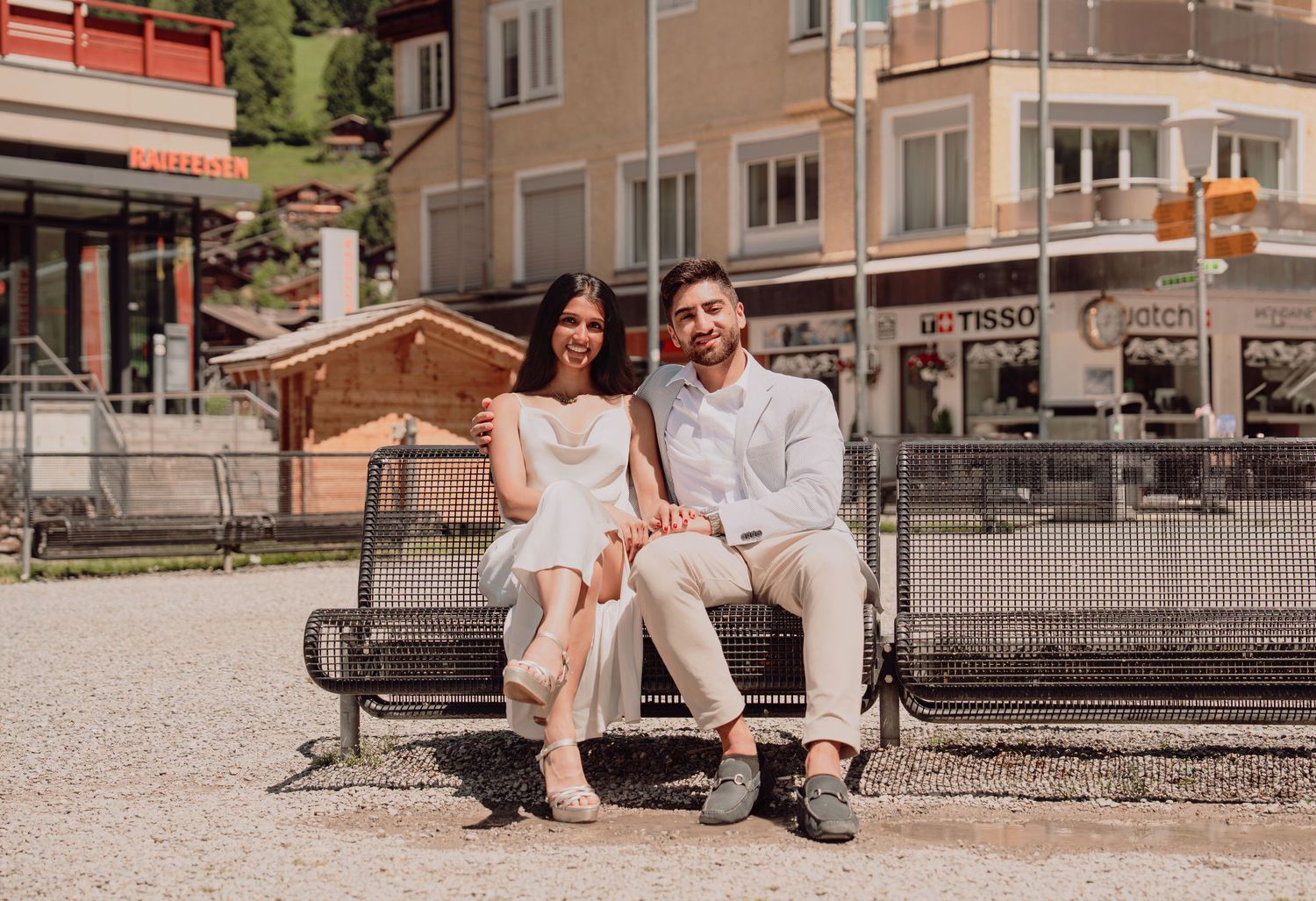 Engaged couple sitting together on a bench in a sunny Swiss Alpine village town square.