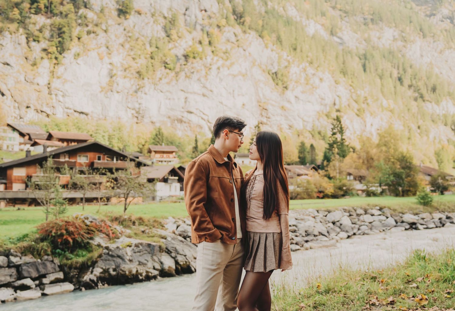 Young Asian couple gazing at each other beside a glacial river with traditional Swiss chalets and autumn foliage.