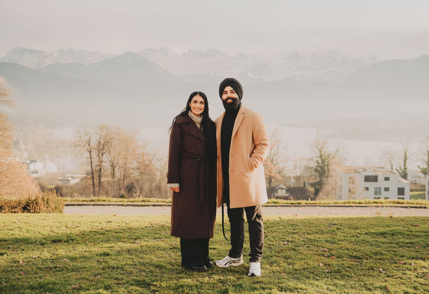 Sikh couple standing together on green lawn with misty mountain cityscape in background.