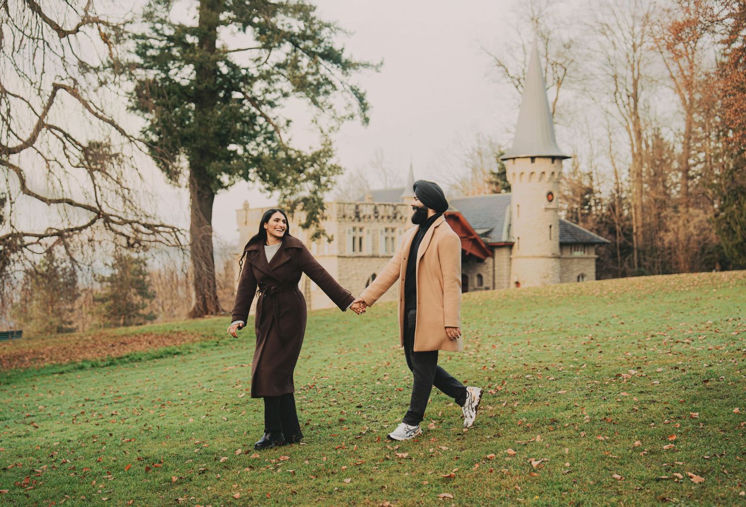Two women holding hands walking through a lush green park with a castle in the background.