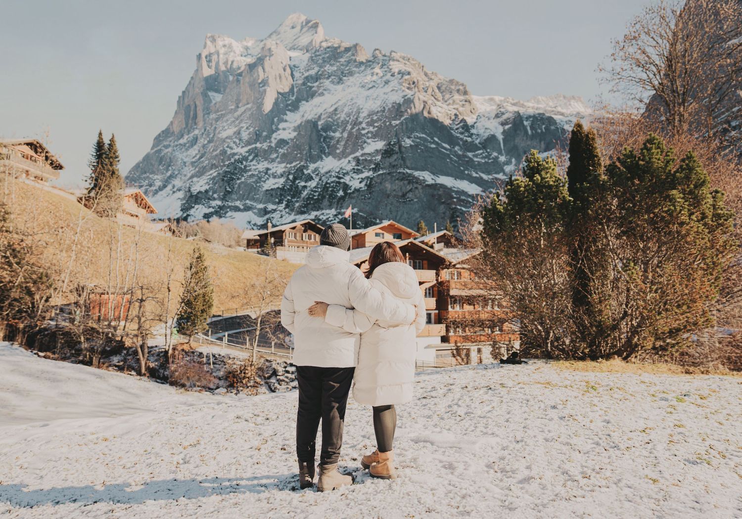 Couple walking arm in arm through snowy alpine village with majestic mountain in background.
