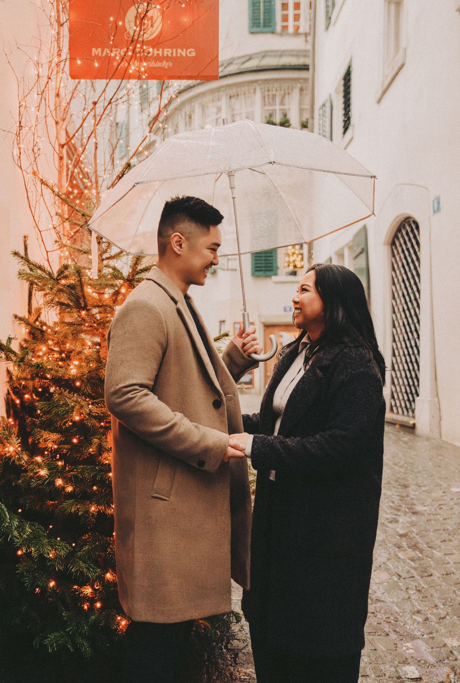 Couple smiling under umbrella beside glowing Christmas tree on rainy street.