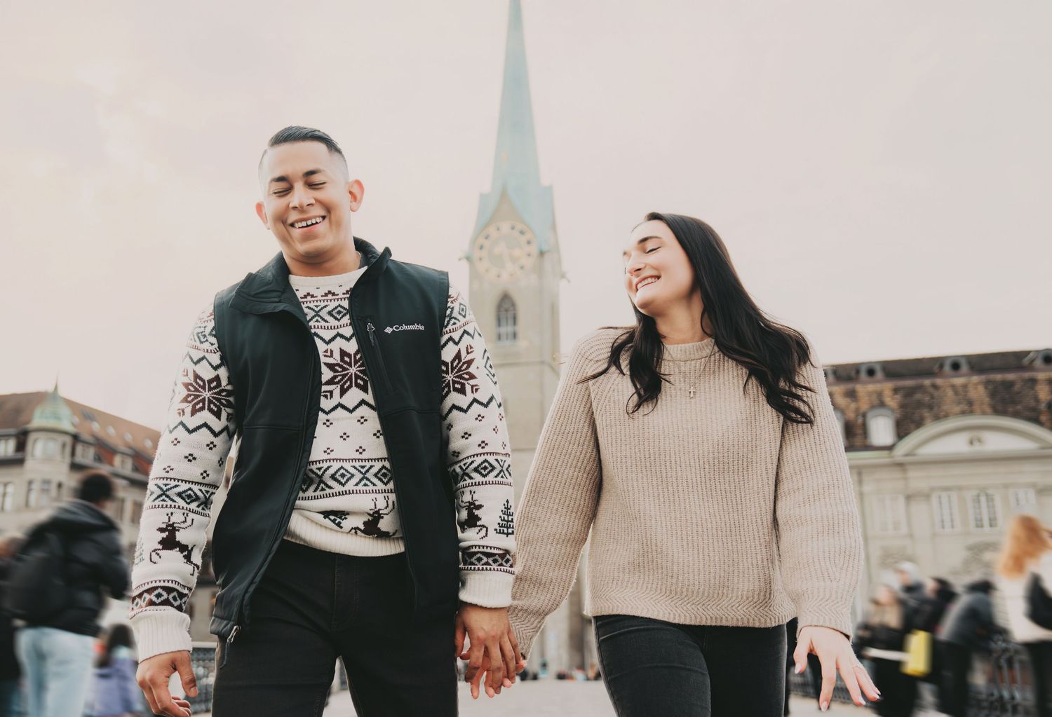 Smiling couple strolling hand in hand with a tall Gothic church spire in the background in Zurich.