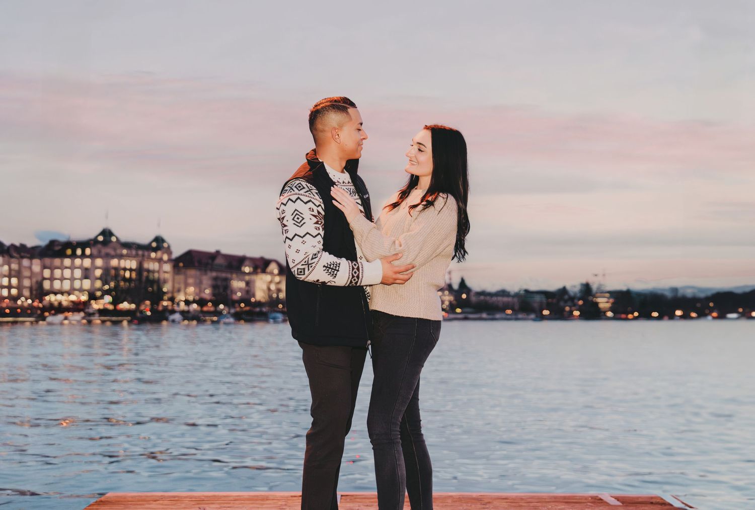 Couple holding each other on a waterfront pier at dusk with city lights behind.